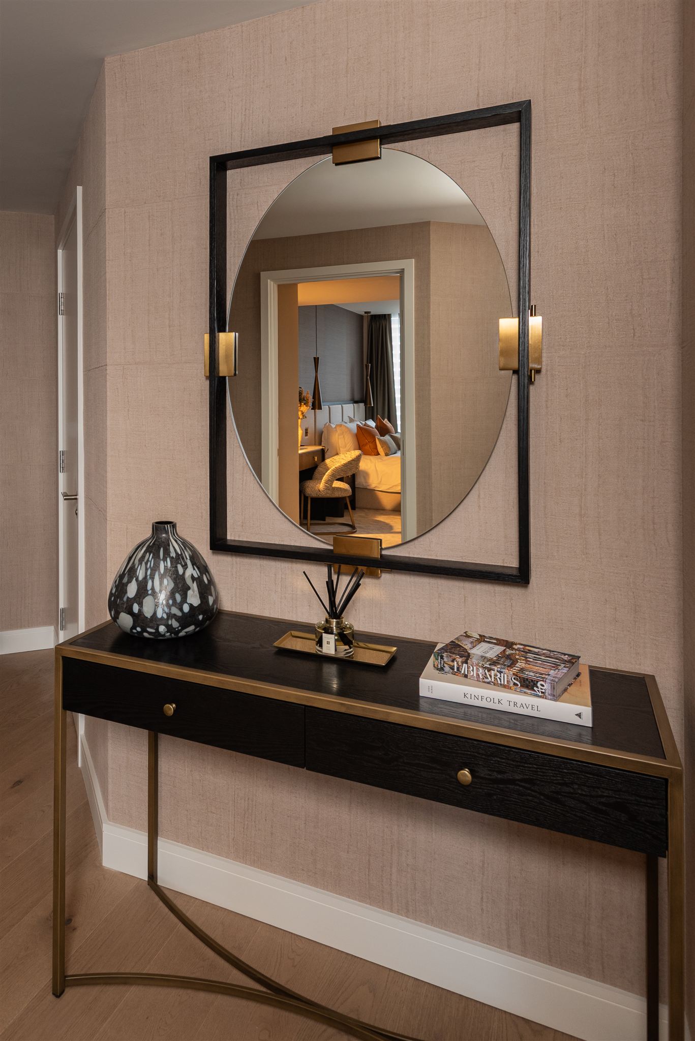 Elegant hallway featuring a black and brass console table beneath a large circular mirror set within a rectangular black frame. Wall sconces with brass detailing flank the mirror. A speckled black vase and lifestyle books including 'Kinfolk Travel' sit on the console surface, with a bedroom visible through the reflection.