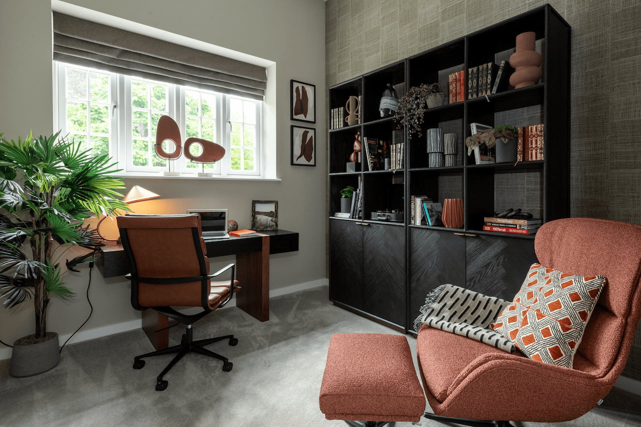 Home office nook with a wooden desk, leather chair, dark built-in bookcase, and a curved terracotta bouclé armchair with ottoman.