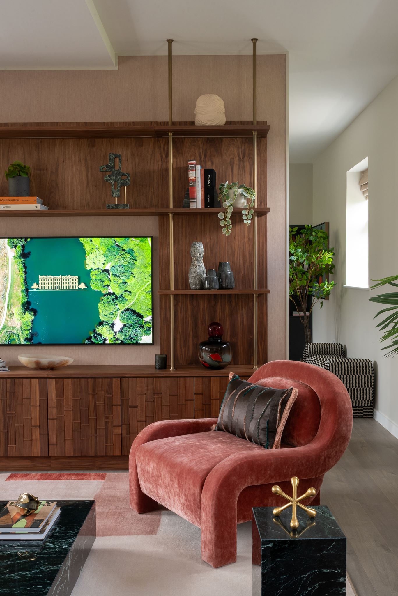 Living room with a walnut shelving wall, curved terracotta velvet armchair, black marble side table, and a wall screen showing an aerial view of the Trent Park estate.