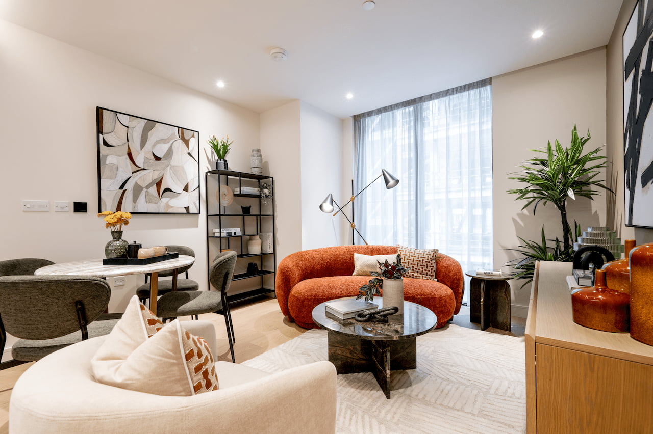 Modern living room with a rust-orange curved sofa with patterned cushions, a black marble coffee table, dark wood shelving styled with books and ceramics, framed gallery-wall art, and a tall potted plant beside floor-to-ceiling windows.