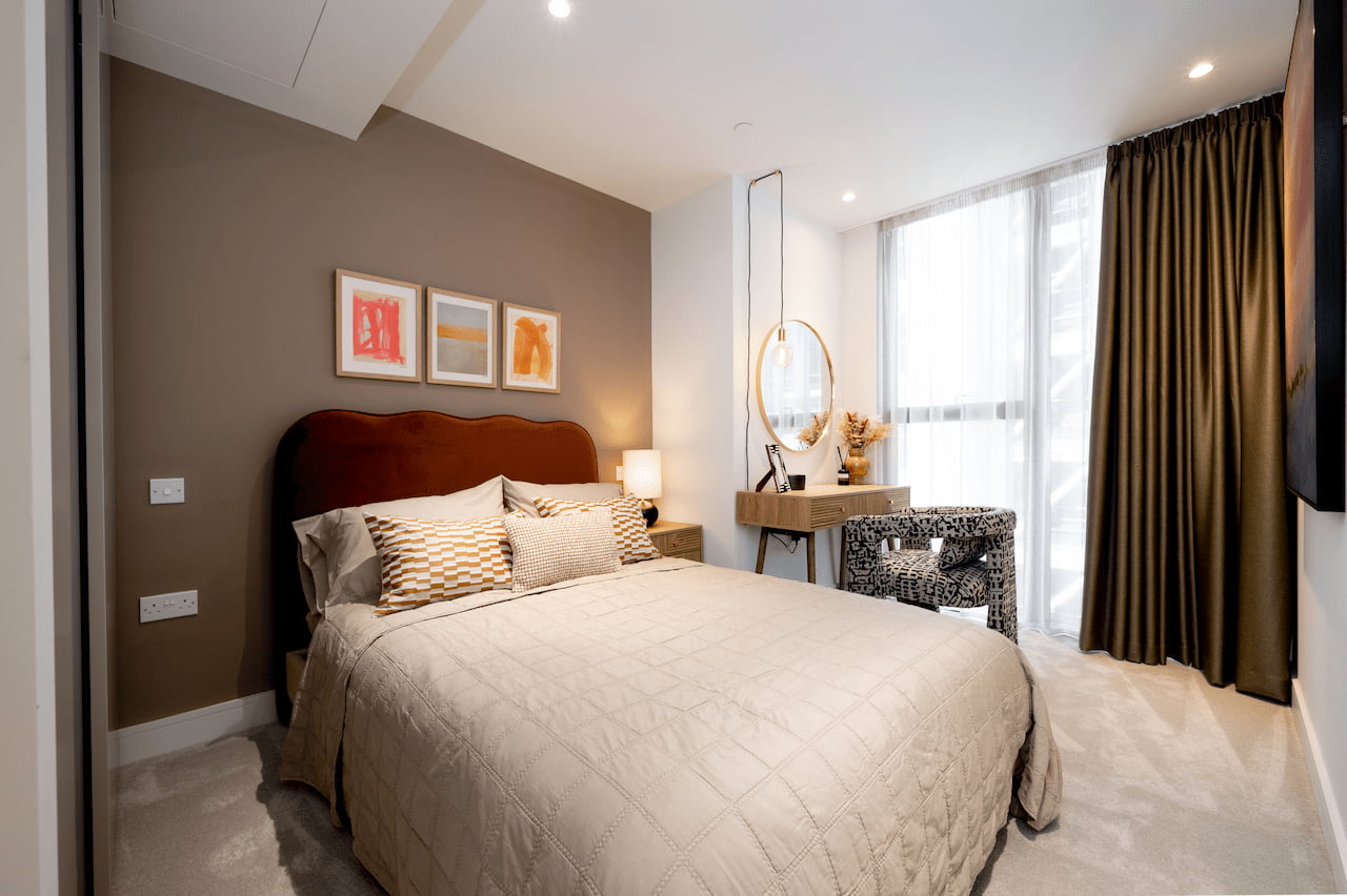 Bedroom with a burgundy wingback headboard, taupe quilted bedding, a mocha feature wall with three small framed prints, a round mirror and vanity table with dried flowers, and olive-green floor-to-ceiling curtains.