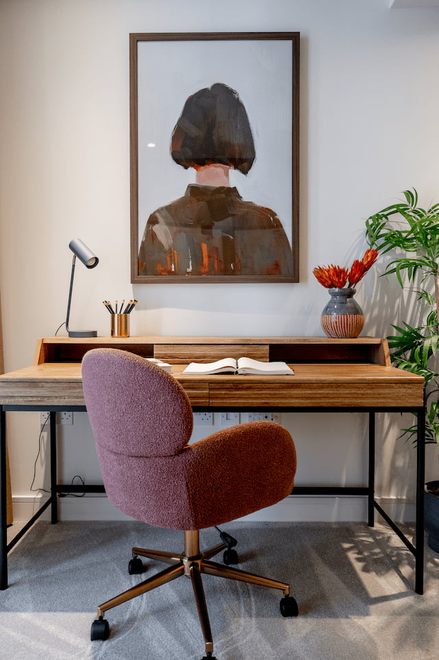 Home office corner with a wooden desk on black metal legs, a mauve boucle swivel chair, an open book, a framed portrait artwork above the desk, and protea flowers in a vase beside a potted plant.