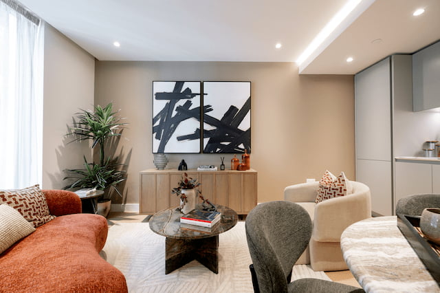 Wide view of the living room with a rust-orange sofa, grey armchairs, a black marble coffee table, a wooden sideboard beneath a large black-and-white abstract artwork, and a potted plant, with the kitchen visible beyond.