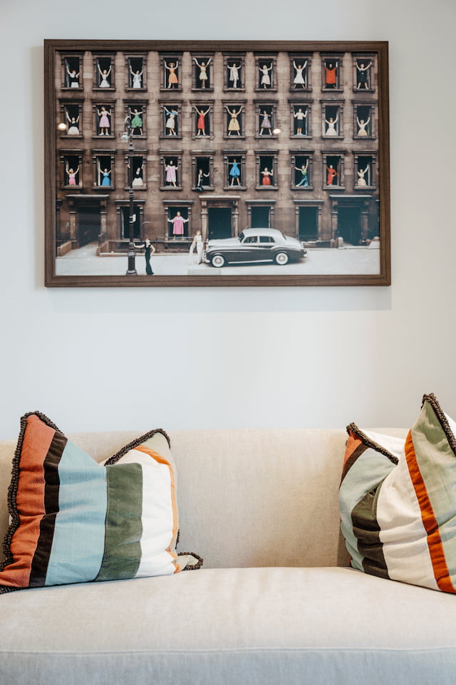 Close-up of a cream sofa with a vintage-style striped scatter cushion, beneath a large framed photographic artwork of a building facade with colourfully dressed figures posing at each window and a classic car parked below.
