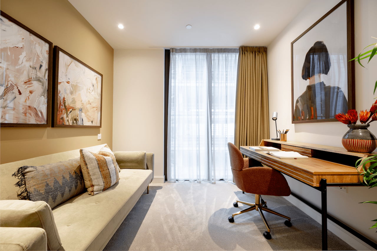Living room and home-office nook with a mustard wall, two abstract framed artworks, a cream sofa with patterned cushions, sheer curtains with amber drapes, and a wooden desk and chair visible on the right.
