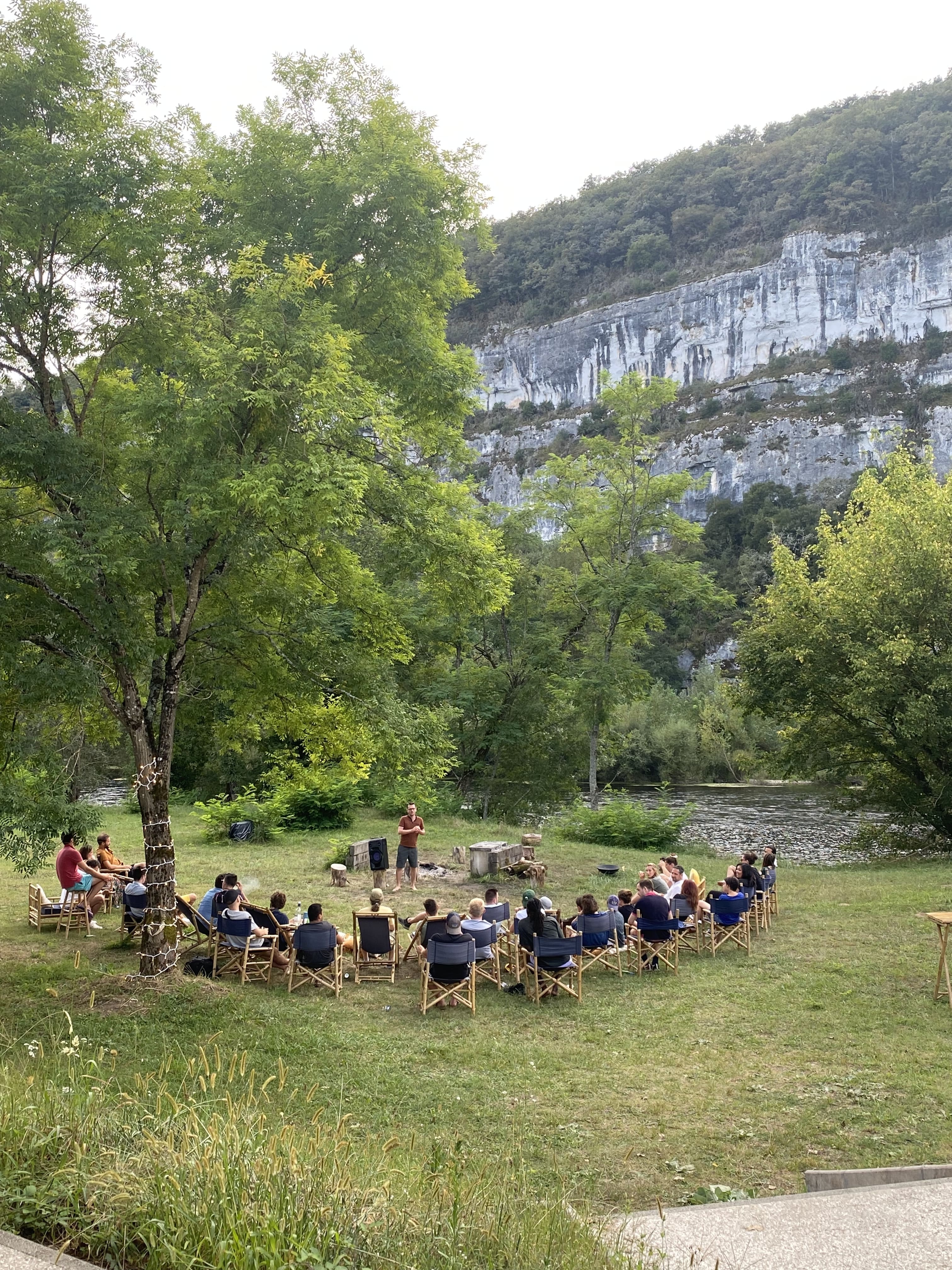 Séminaire d’entreprise en plein air, un groupe de participants assis en cercle écoute un intervenant au bord d’une rivière, entouré de verdure et falaises.