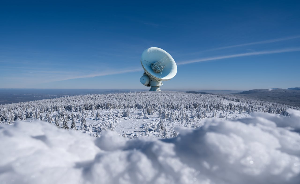 Grande antenne parabolique installée au sommet d’une montagne enneigée sous un ciel bleu.