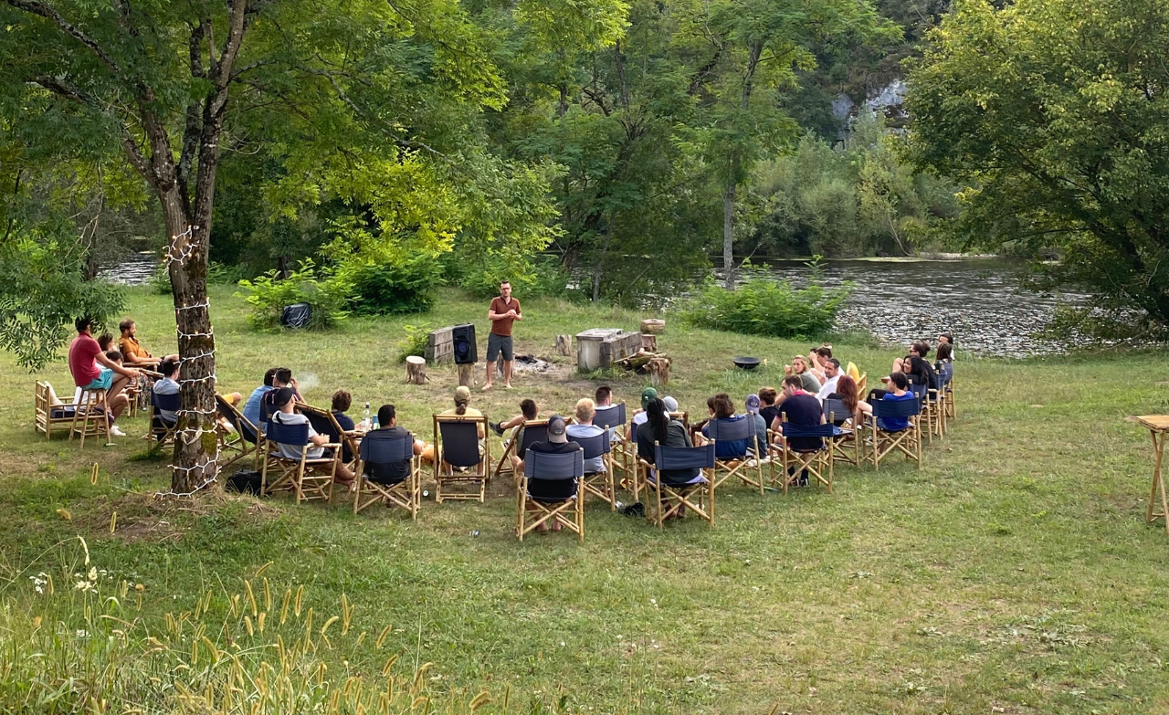 Séminaire en plein air avec un groupe de participants assis en cercle, écoutant un intervenant au bord d’une rivière.
