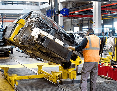 Ouvrier en gilet orange manipulant un véhicule sur un support mécanique dans une usine automobile.
