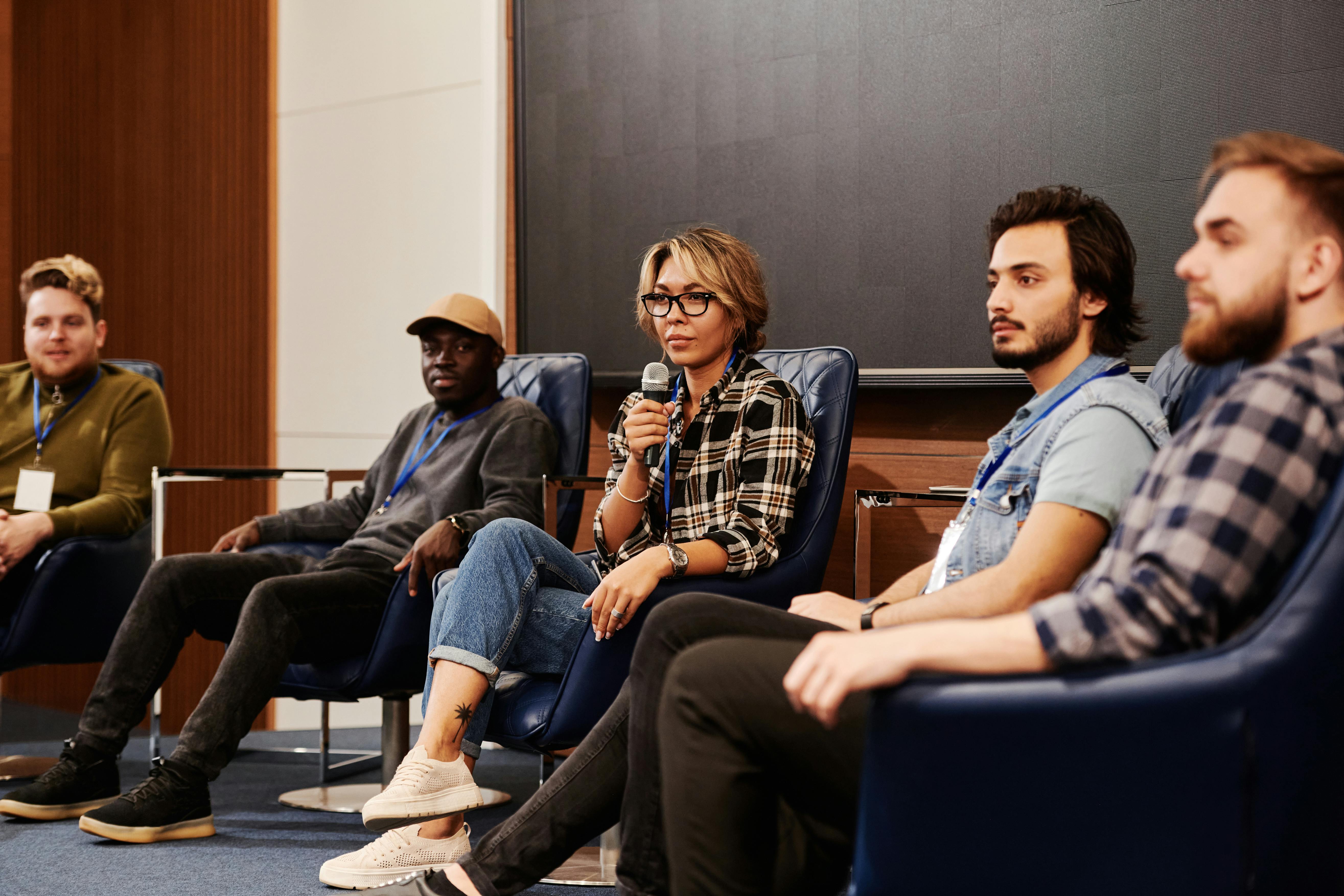 Panel of five diverse people seated on stage chairs, with the woman in the center holding a microphone during a discussion.