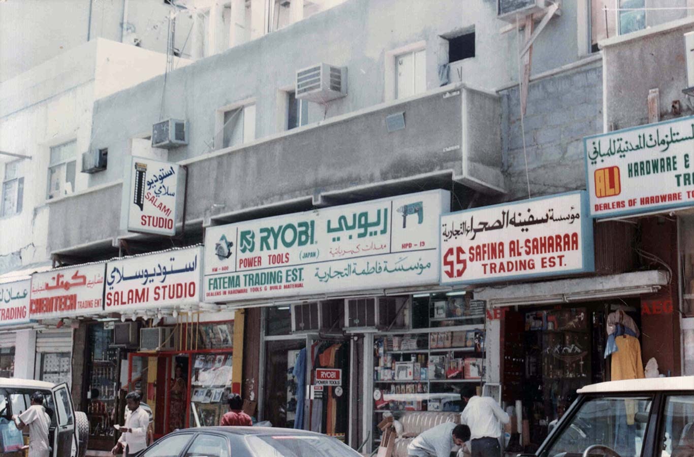 Street view of shops with signs for Salami Studio, Ryobi Power Tools, Fatema Trading Est., and Safina Al-Saharaa Trading Est., with people and cars in front.