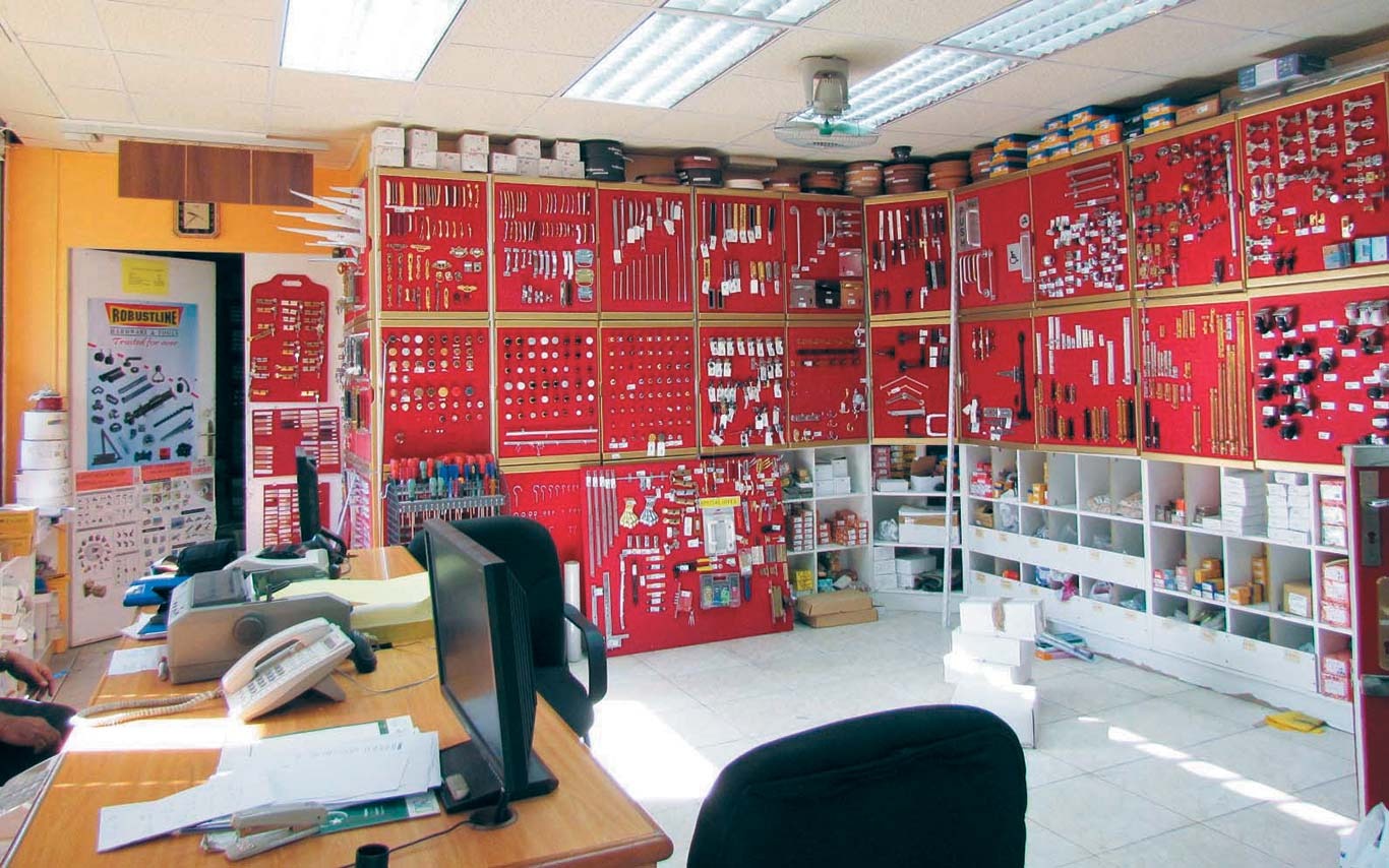Hardware store showroom with red display boards showcasing various tools and fittings, and a wooden desk with office equipment.