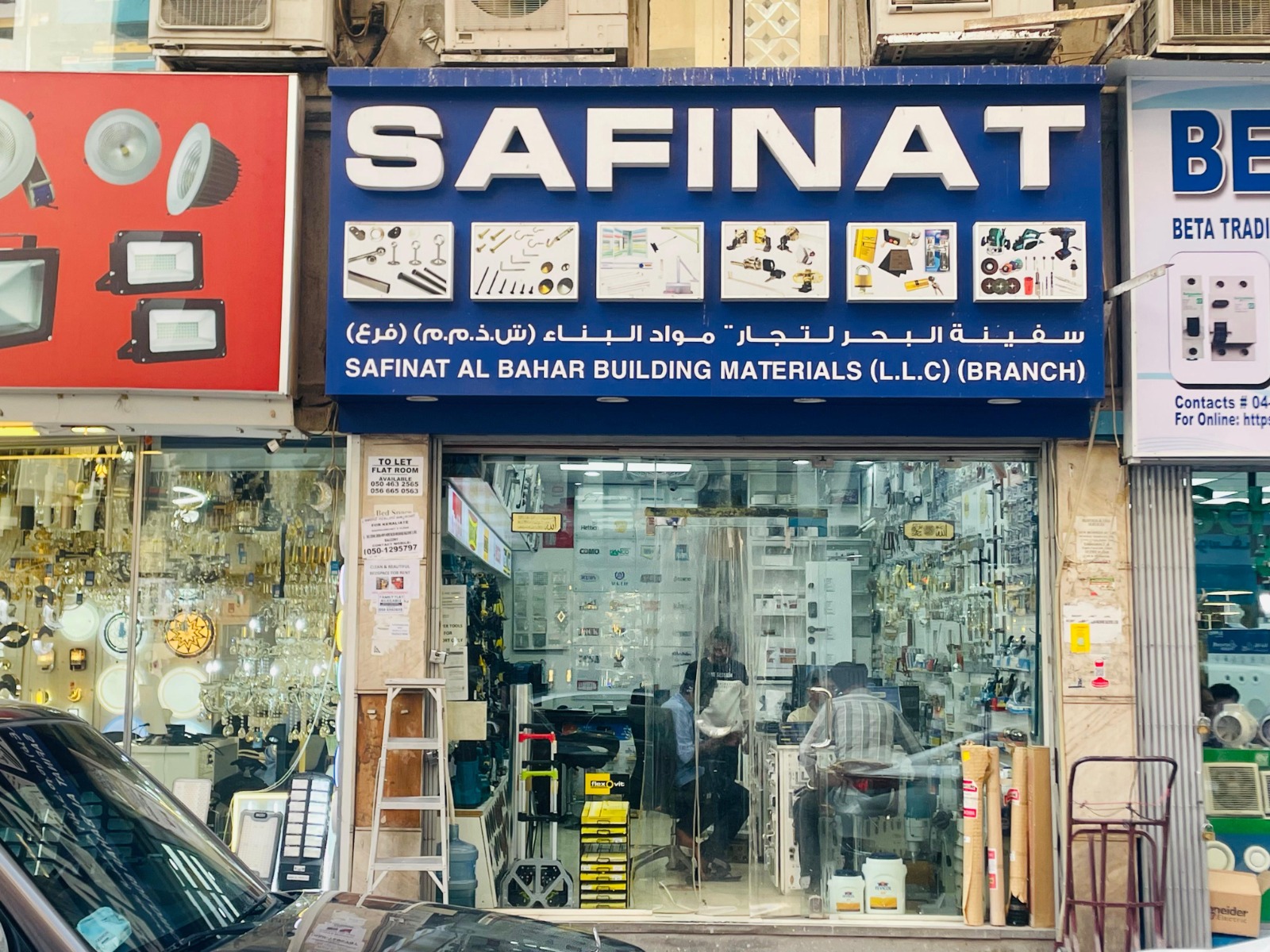 Shopfront of Safinat Al Bahar Building Materials showing a blue signboard with images of hardware tools and materials above a glass entrance with people inside.