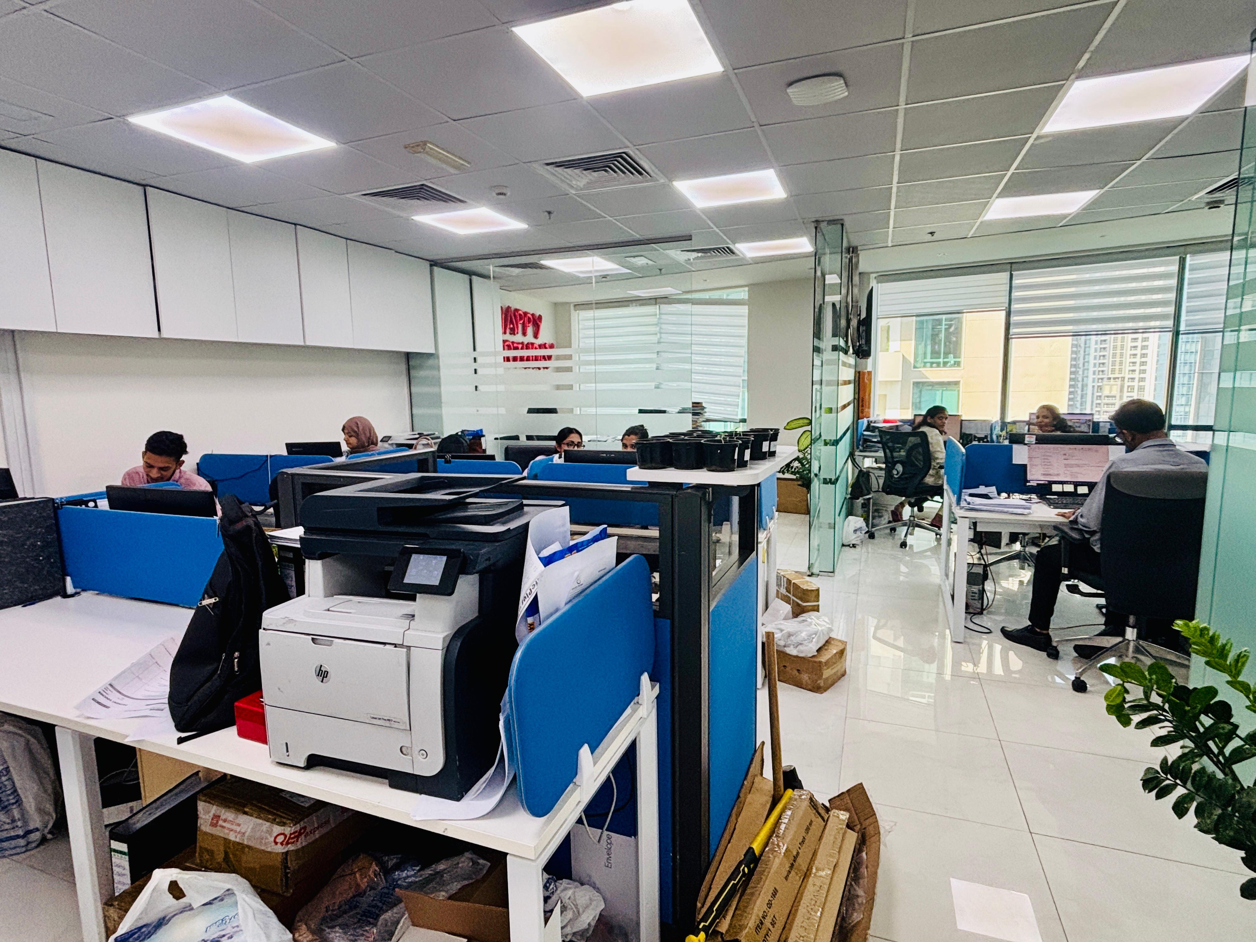 Office space with several employees working at desks with blue partitions, a printer on a table, and large windows with city views.