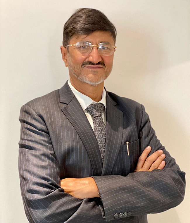 Man with glasses wearing a gray pinstripe suit and patterned tie, standing with arms crossed against a plain light background.