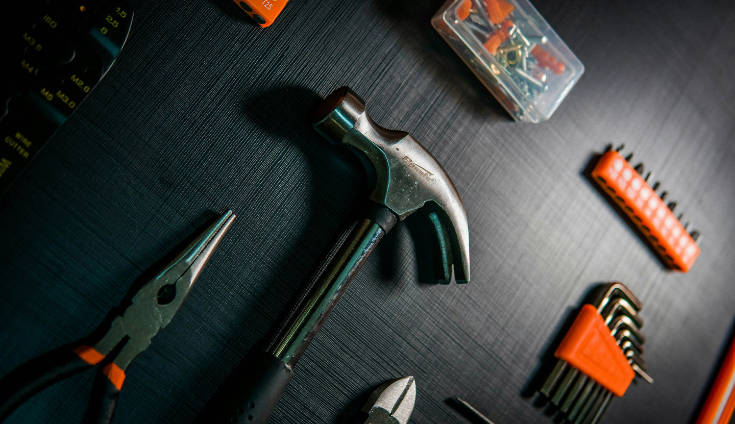 Close-up of a claw hammer, needle-nose pliers, wire cutter, hex keys, screwdriver bits, and small screws on a dark textured surface.