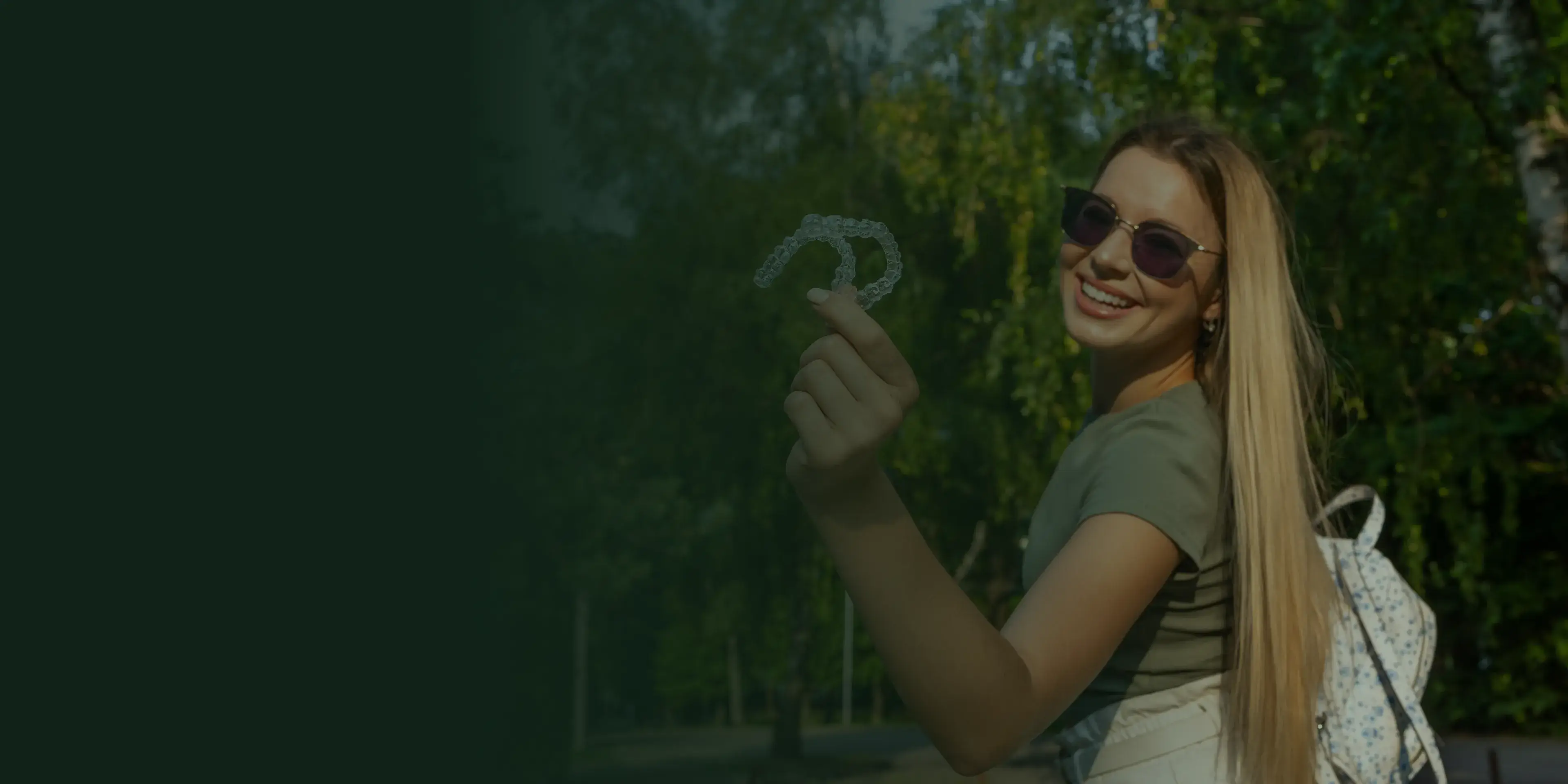 A smiling woman wearing sunglasses holds up a dental retainer outdoors.