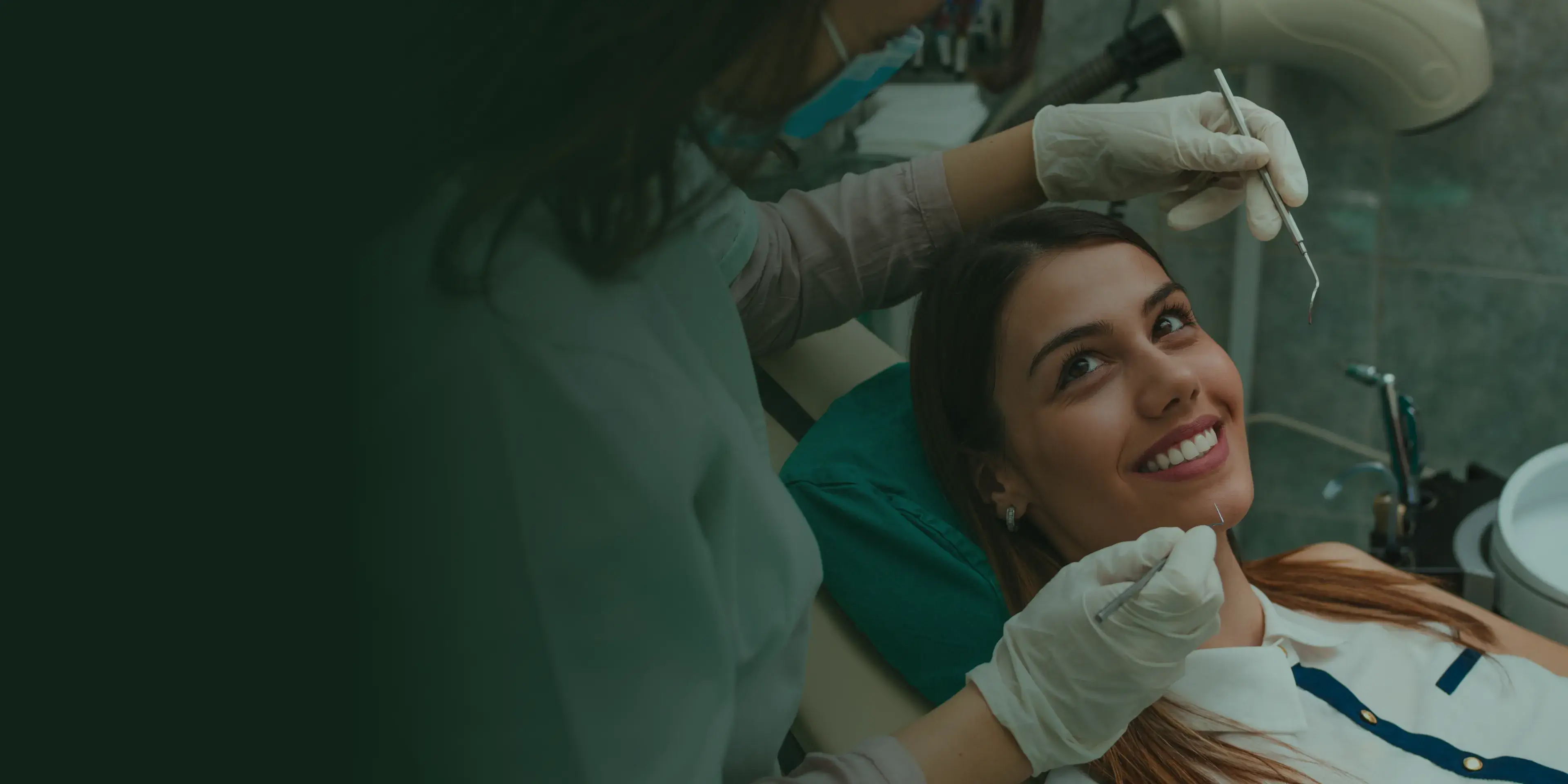 A person smiling warmly, clasping hands together, standing outside near a tree and a brick wall.