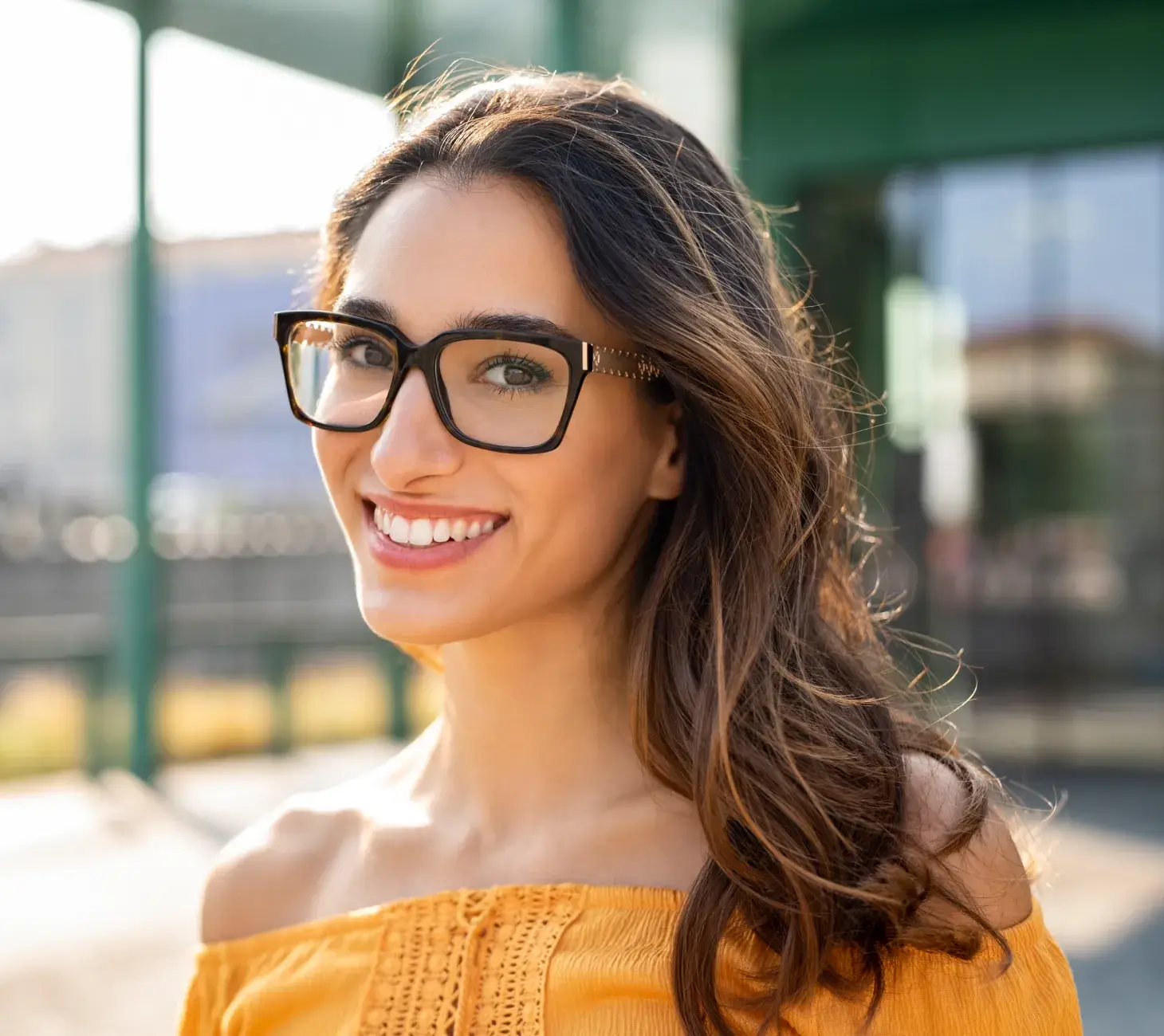 A woman with long hair and glasses smiles while standing outside.