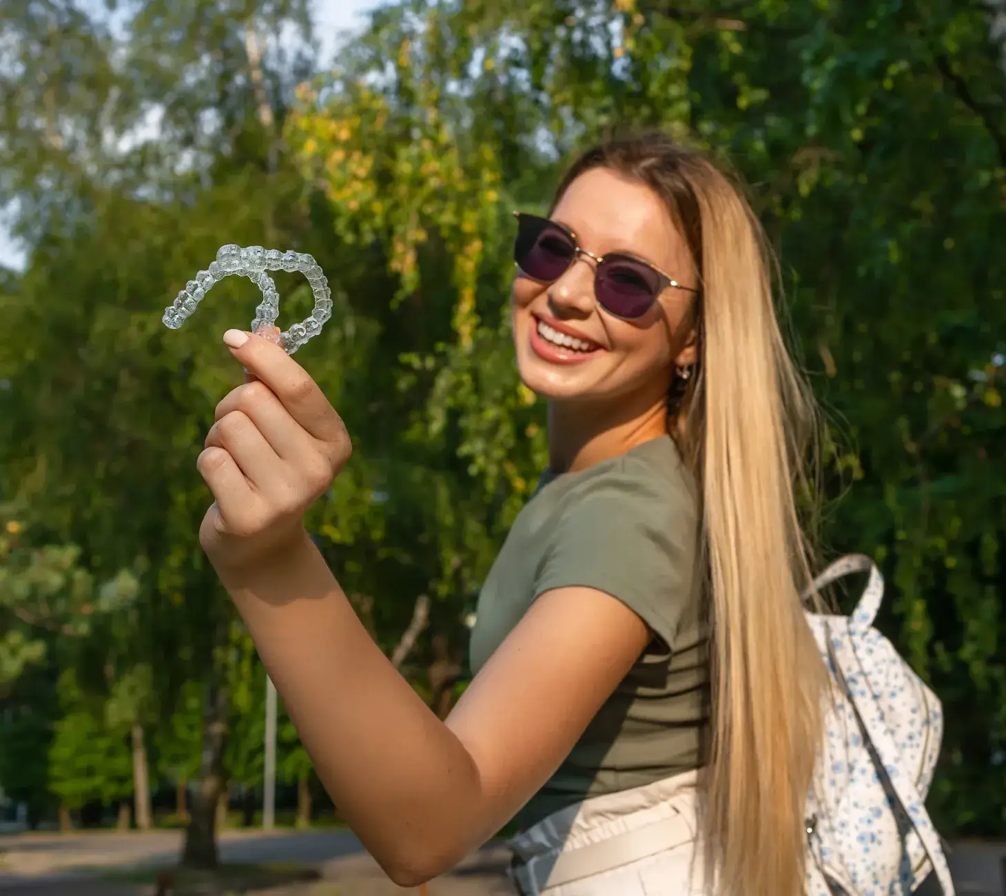 A woman smiling and holding up a pair of clear aligners outdoors.