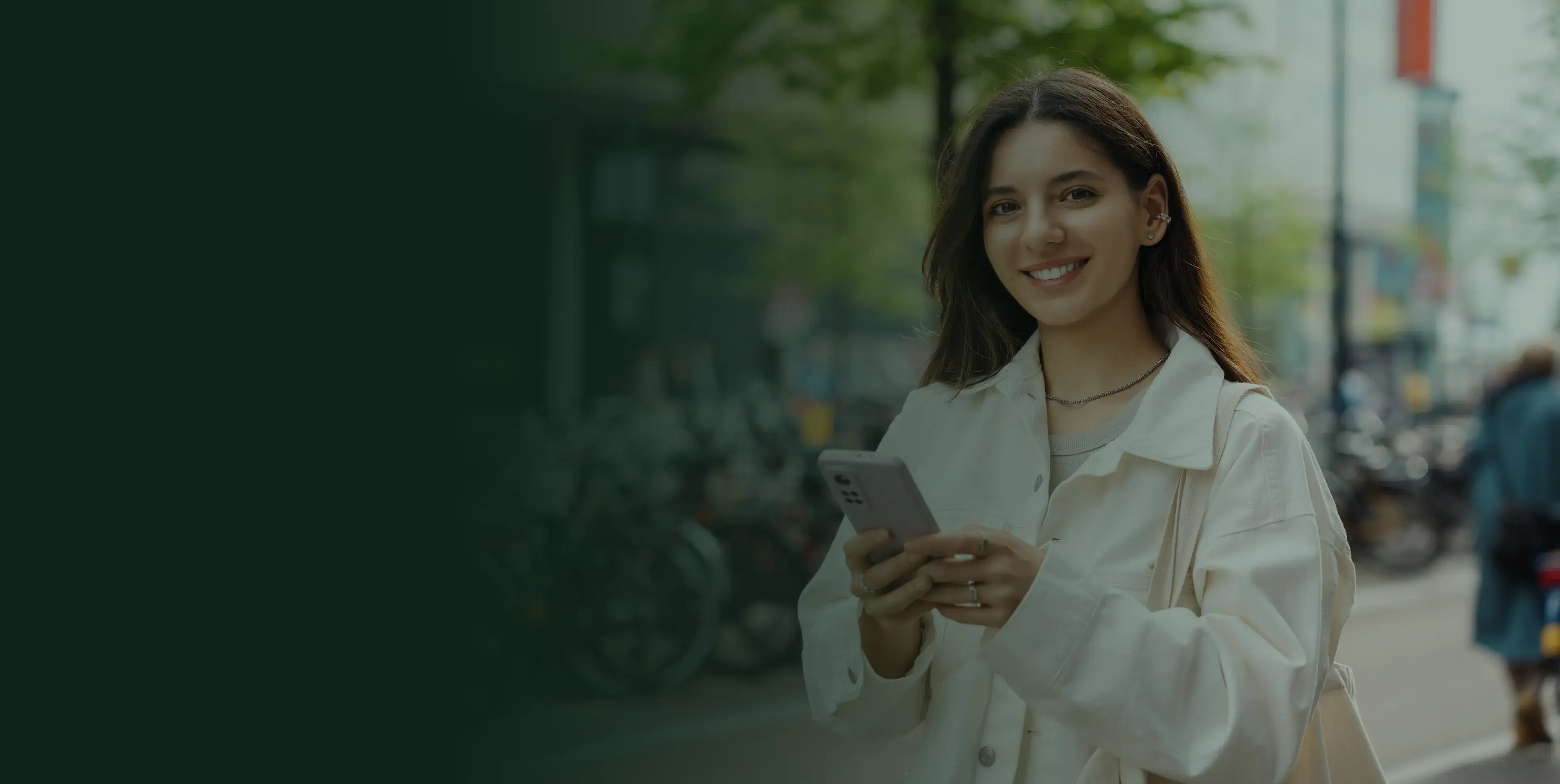 A woman smiles while holding a smartphone, standing outside with trees in the background.