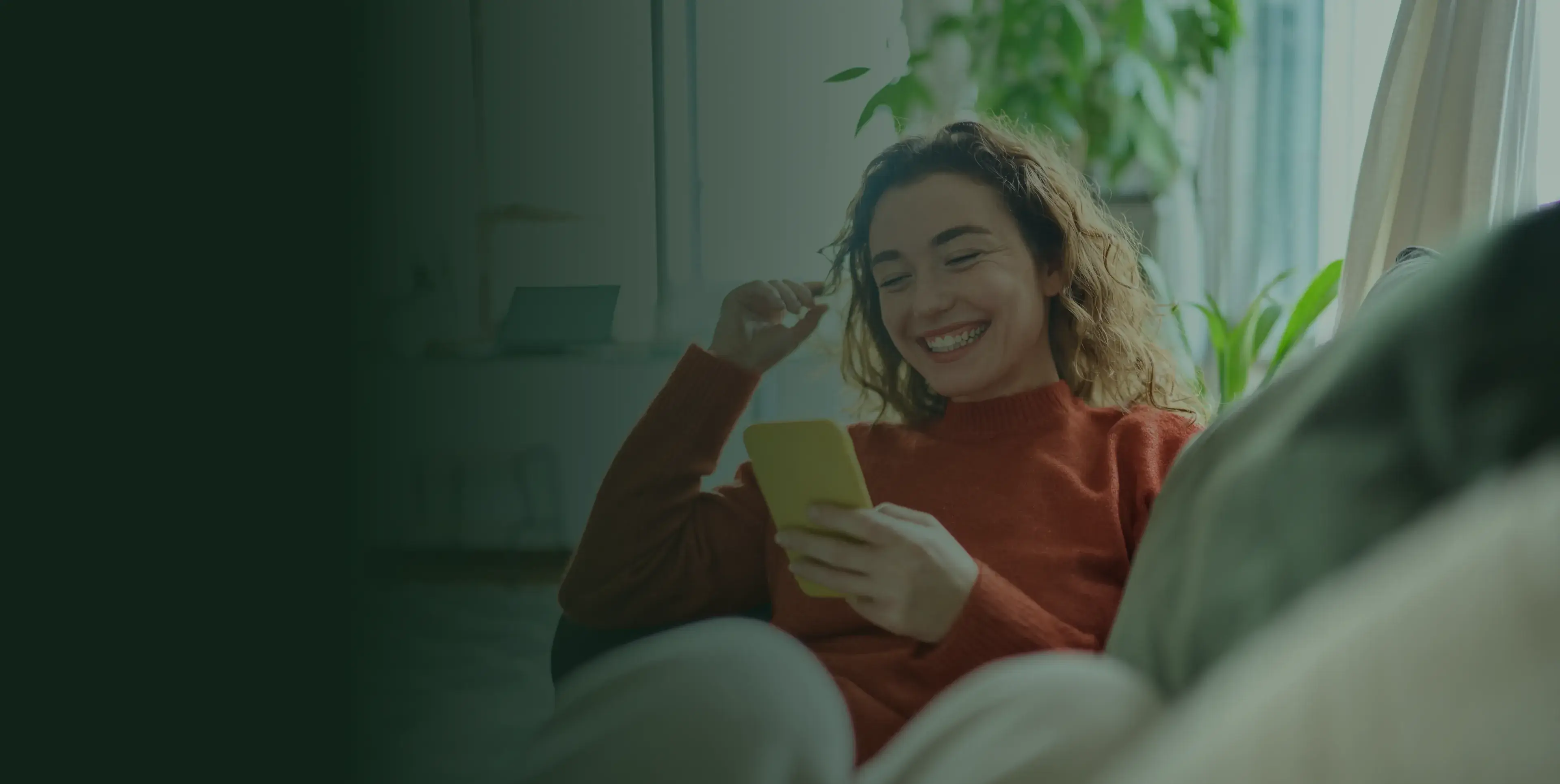 A woman smiles while looking at her phone, sitting on a couch indoors.