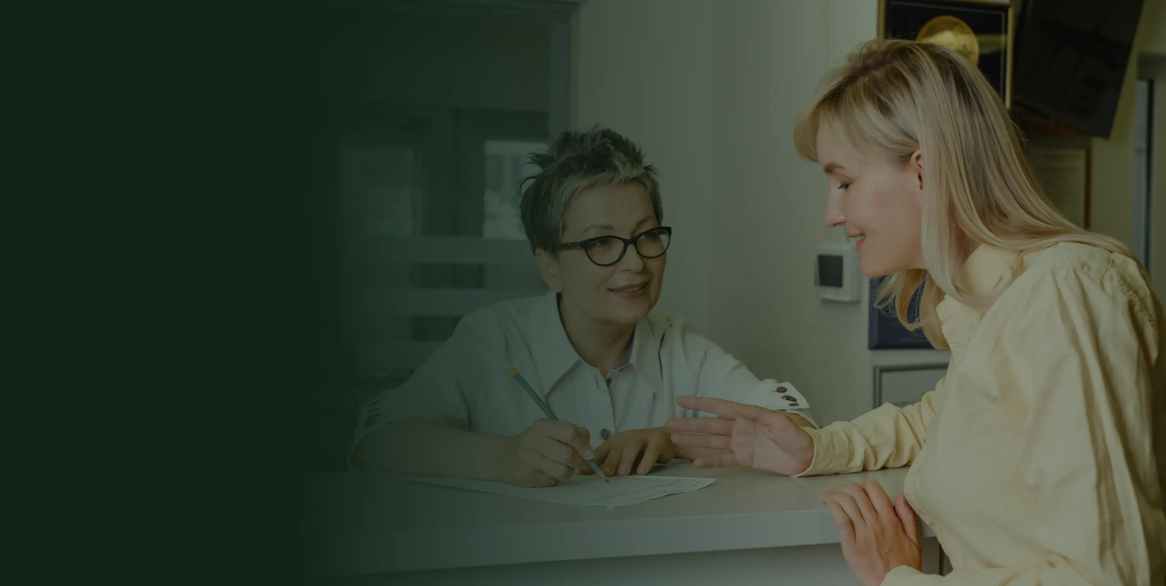 A woman in a yellow shirt talks to a receptionist at a desk, holding a pen.