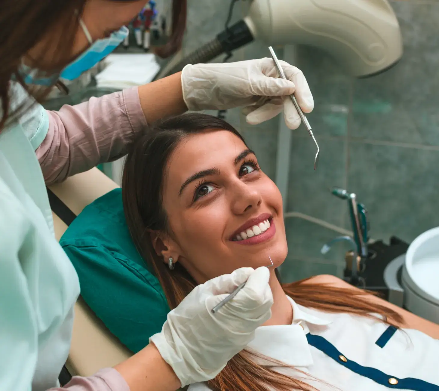 A dentist is examining a smiling woman's teeth using dental tools in a clinic.