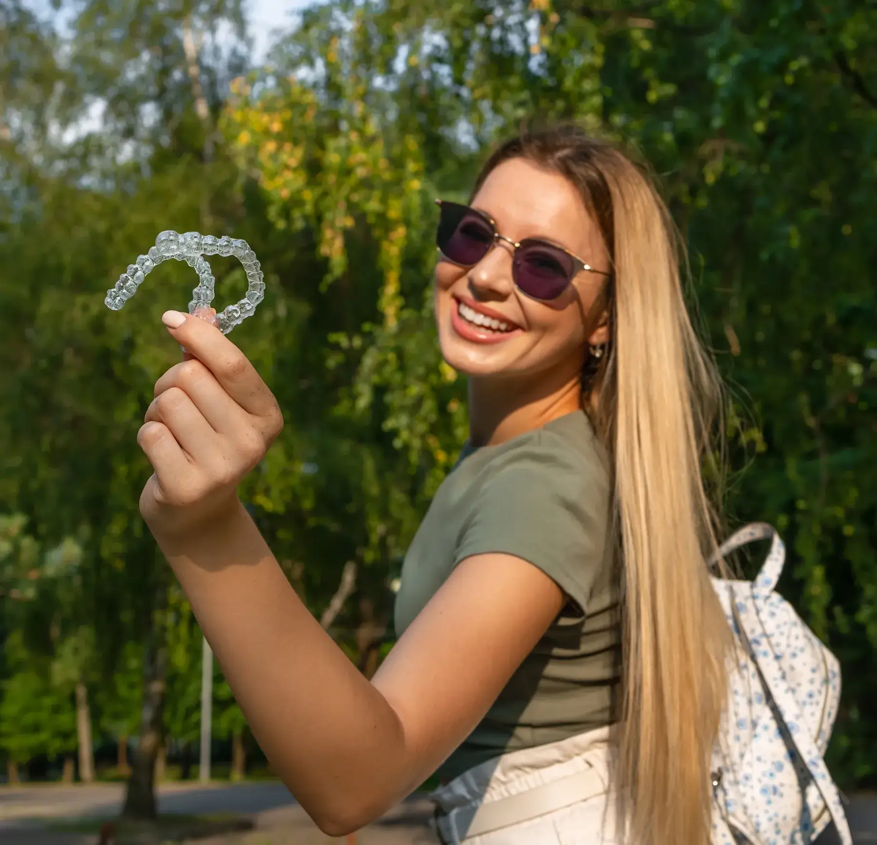 A woman with long hair and sunglasses smiles while holding a clear dental aligner outdoors.