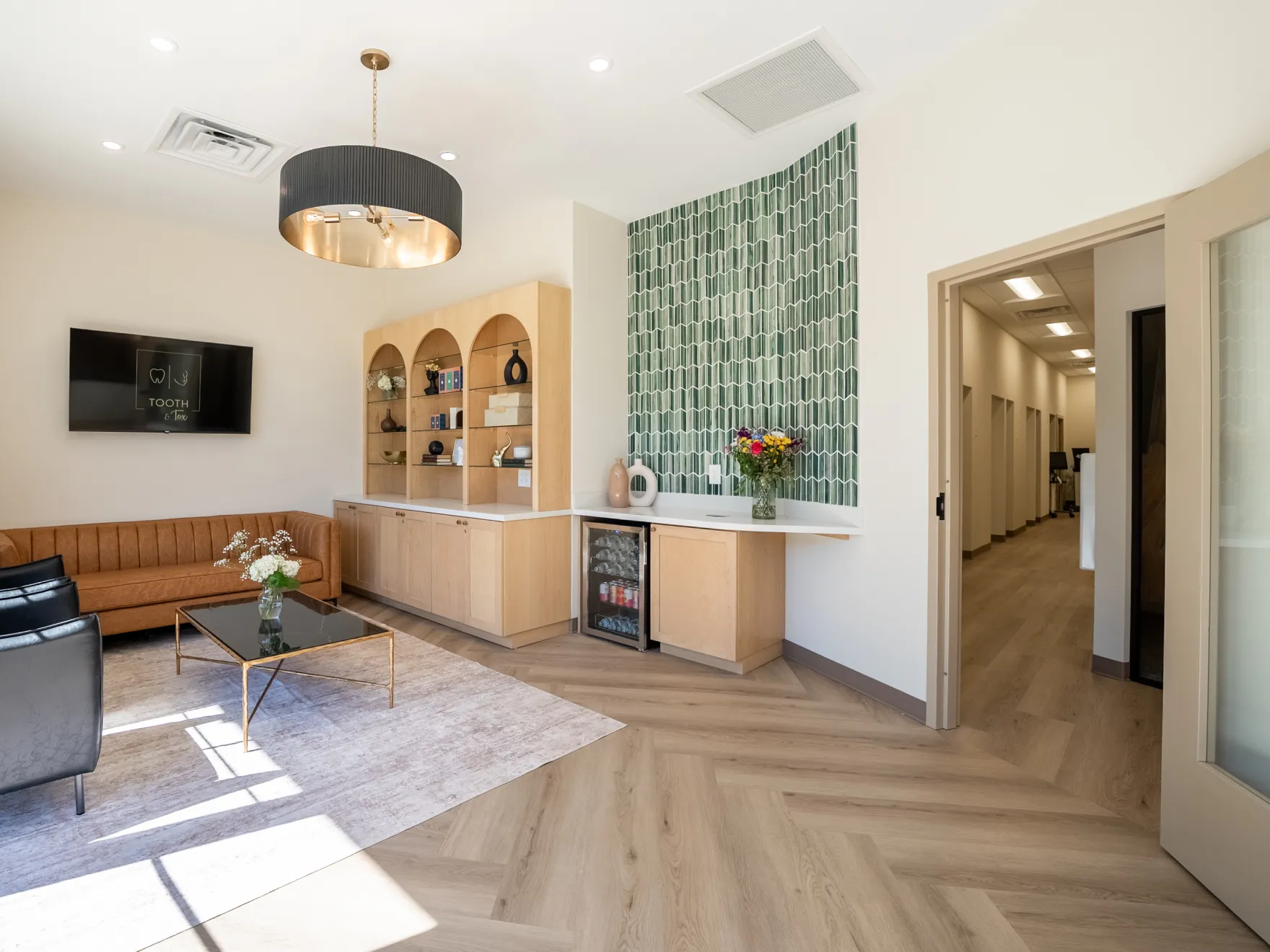 Modern waiting room with a leather sofa, armchairs, TV, shelves, and flowers on a countertop.