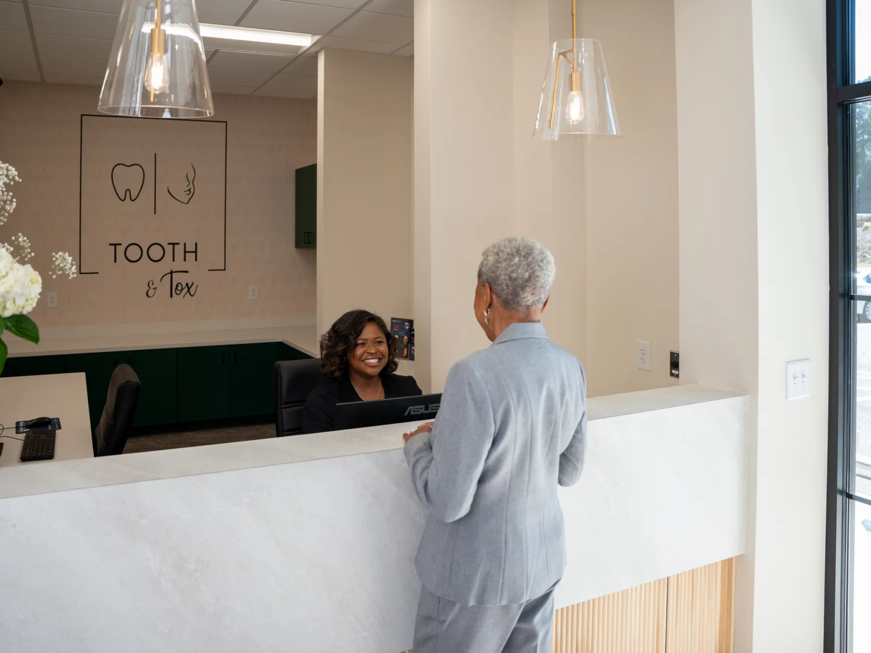 A woman in a gray suit talks to a receptionist at a dental office counter.