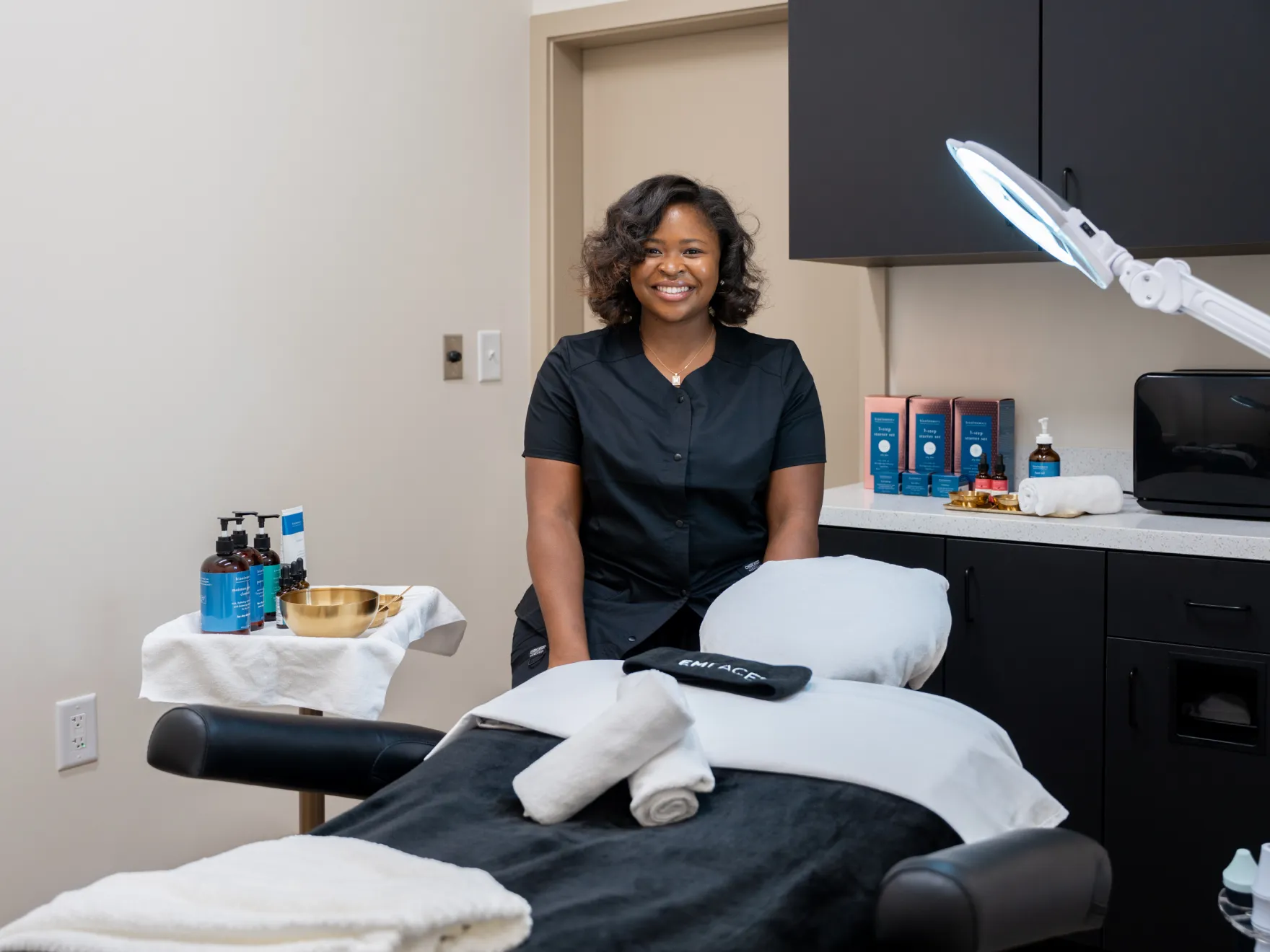 A woman in a black uniform stands smiling beside a treatment bed in a spa room.