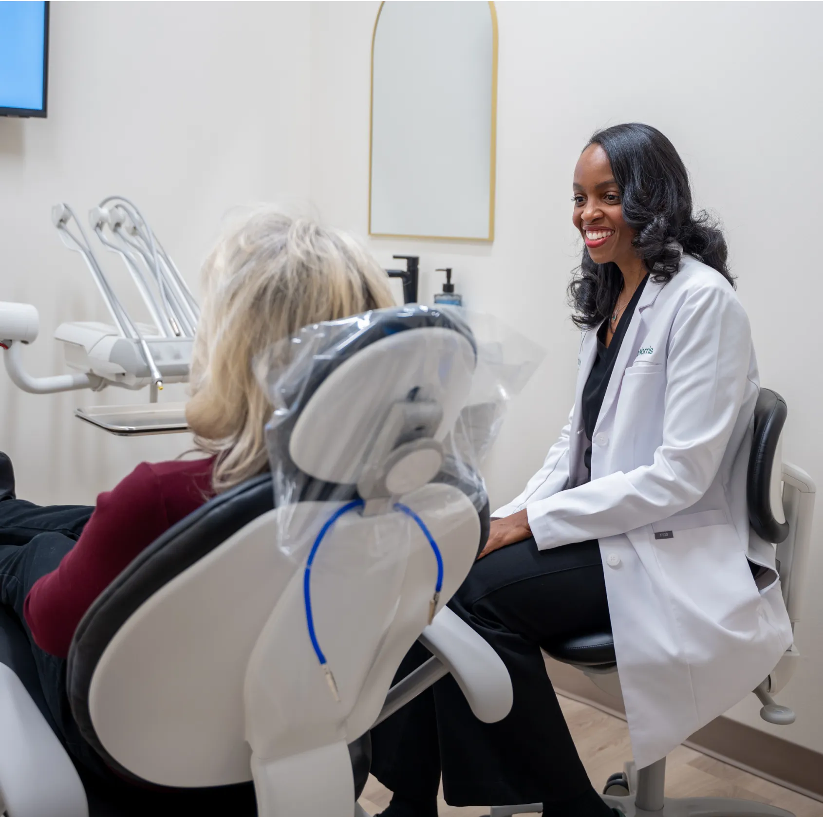 A dentist in a white coat talks to a patient sitting in a dental chair.