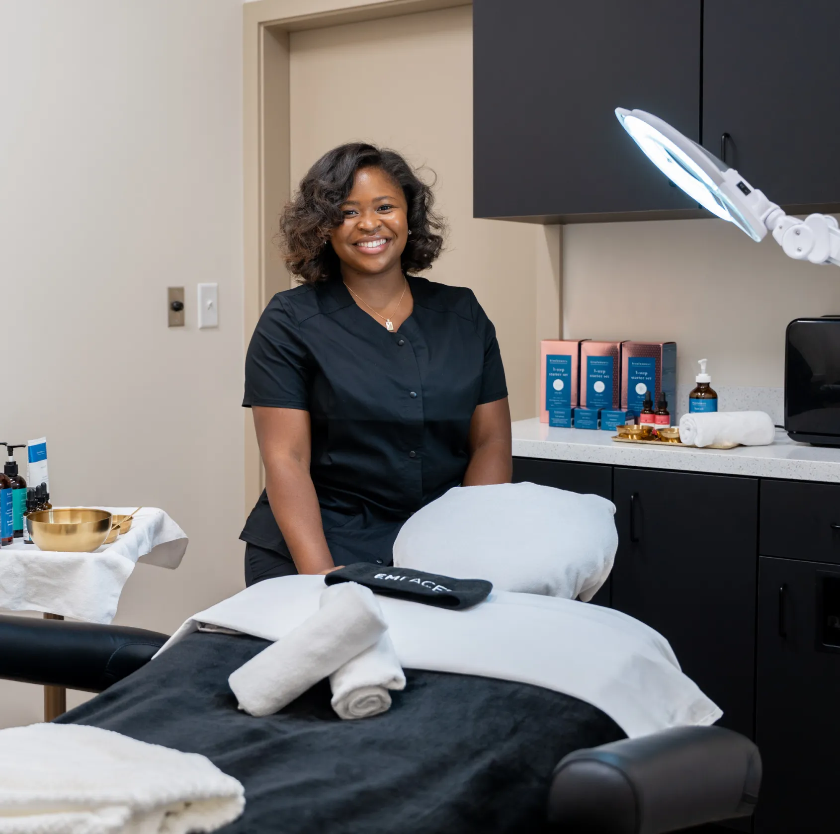 Woman smiling behind a treatment table in a spa, with skincare products on the counter.
