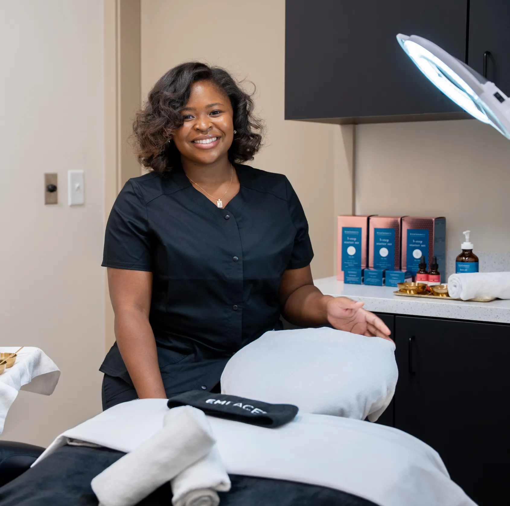 A woman in a black uniform smiles while standing in a spa treatment room.