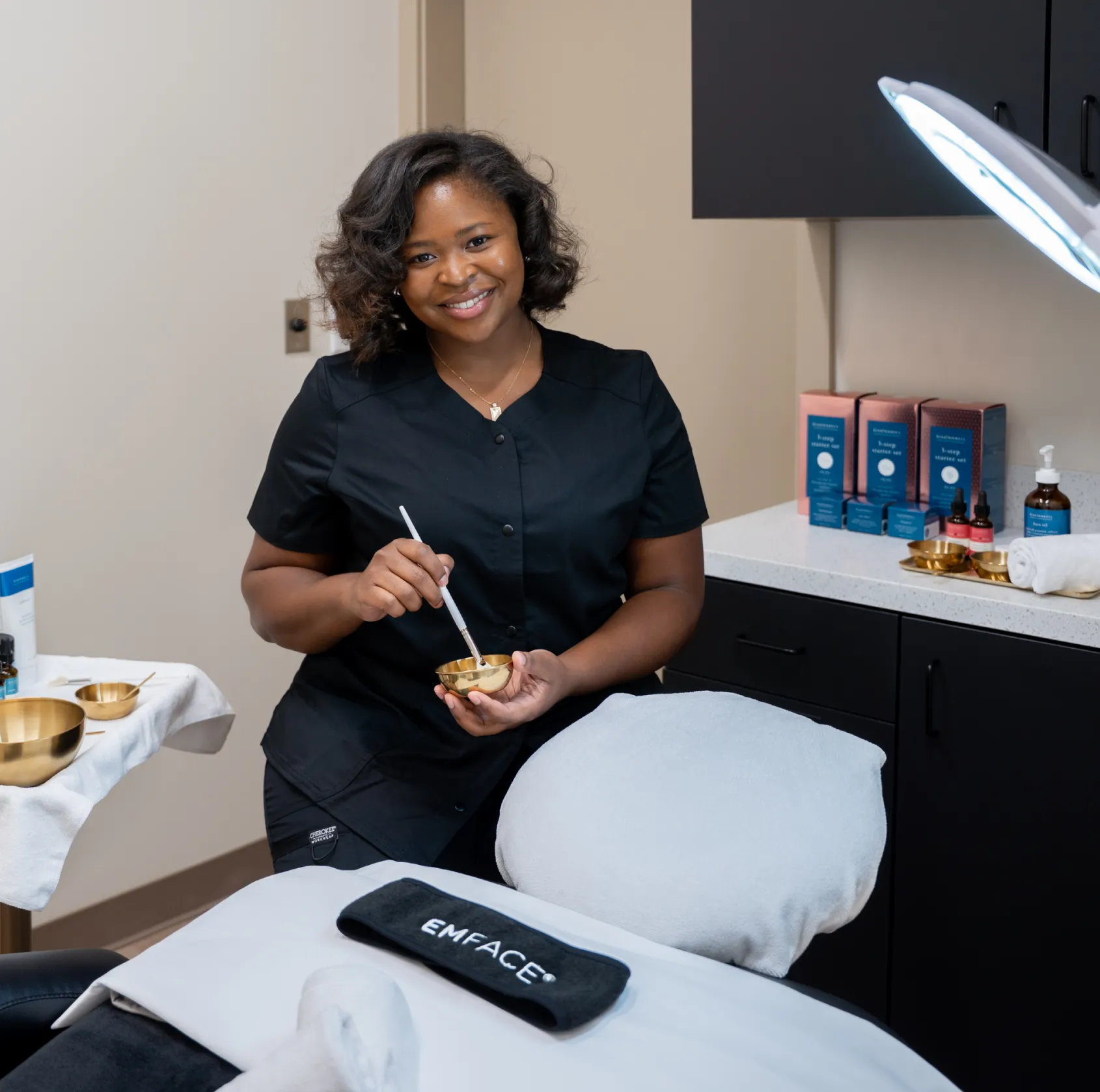 A woman in scrubs stands in a treatment room, holding a small bowl and brush.
