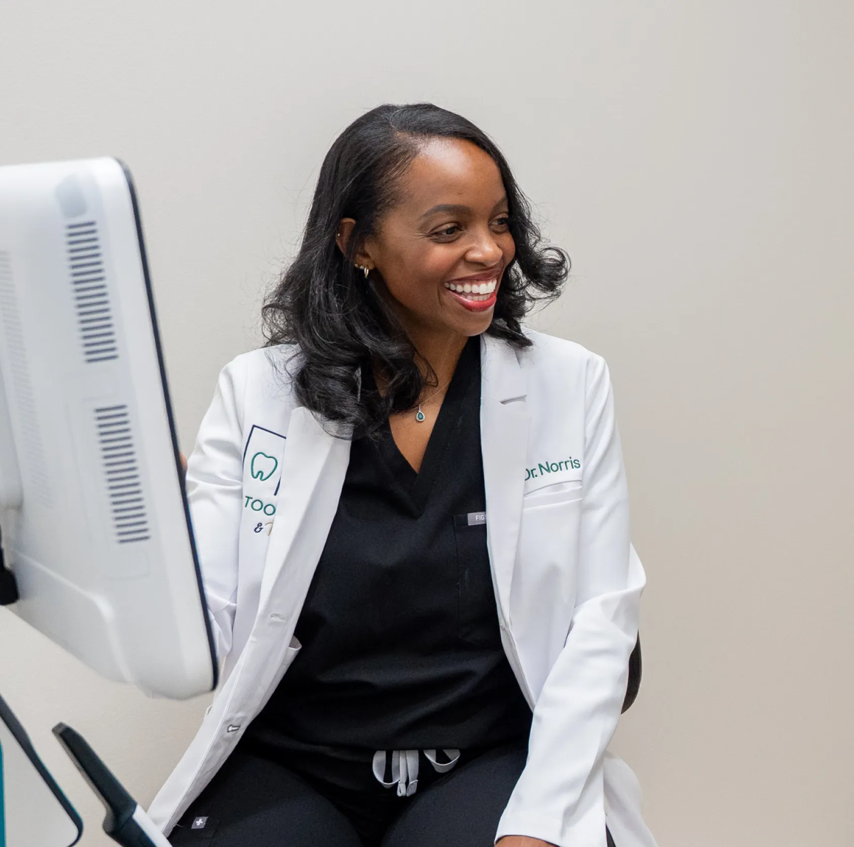 A smiling woman in a lab coat sits near a computer monitor.