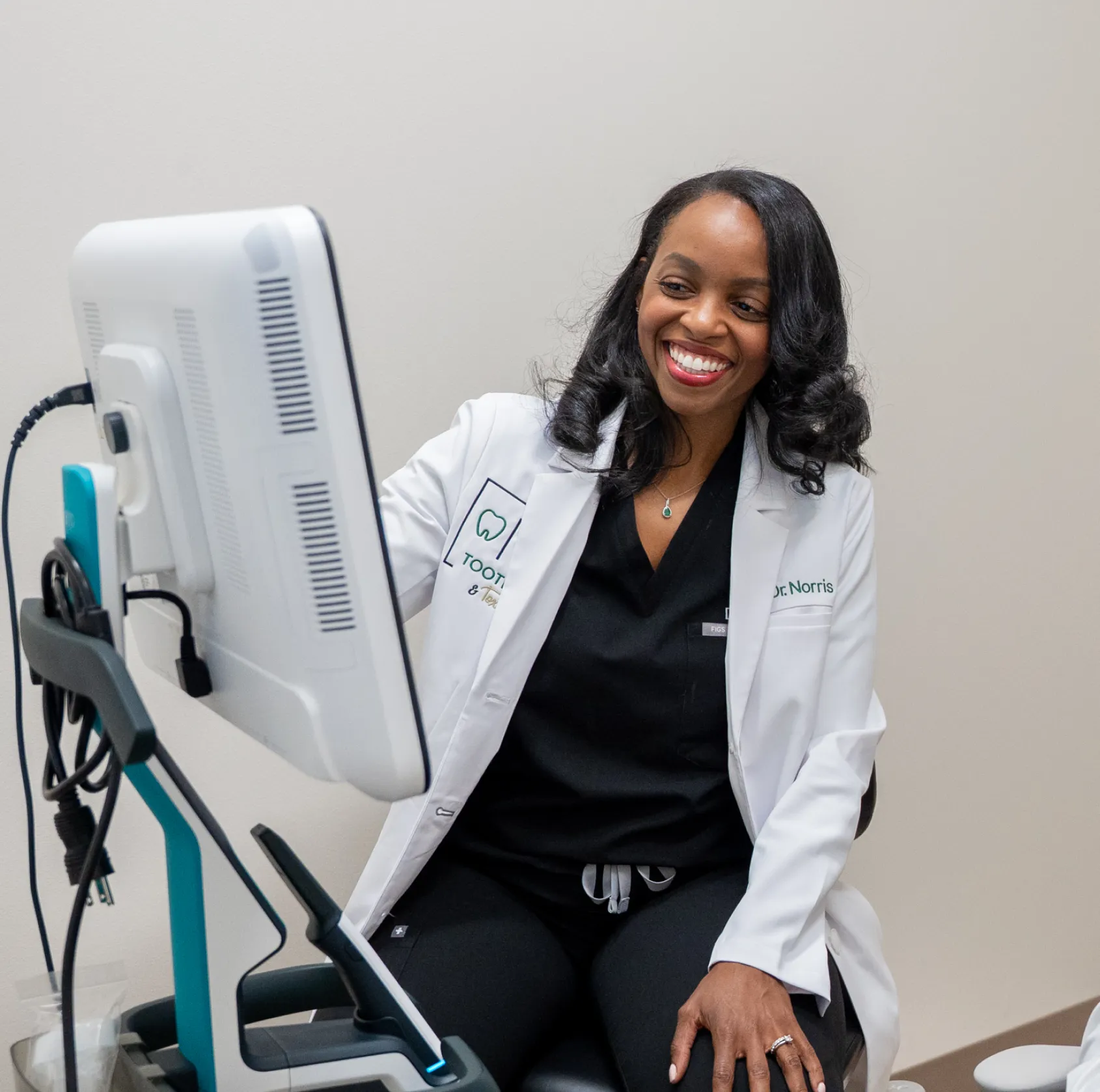 A dentist in a white coat smiles while looking at a computer screen in an office.