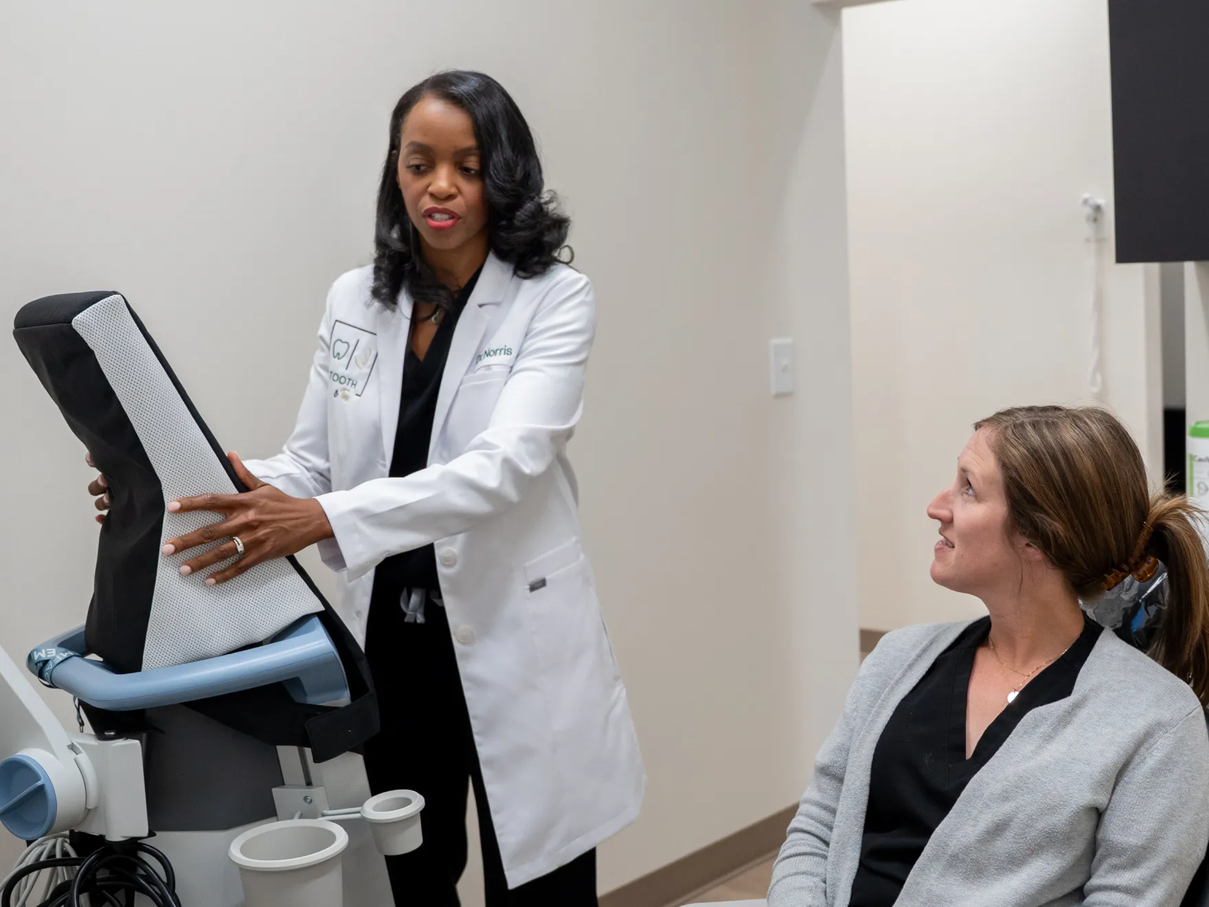 A doctor in a lab coat explains a medical device to a seated woman in an exam room.