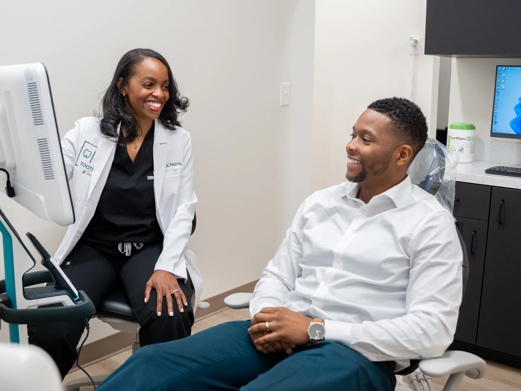 A dentist and patient talk cheerfully in a dental office with a computer in the background.