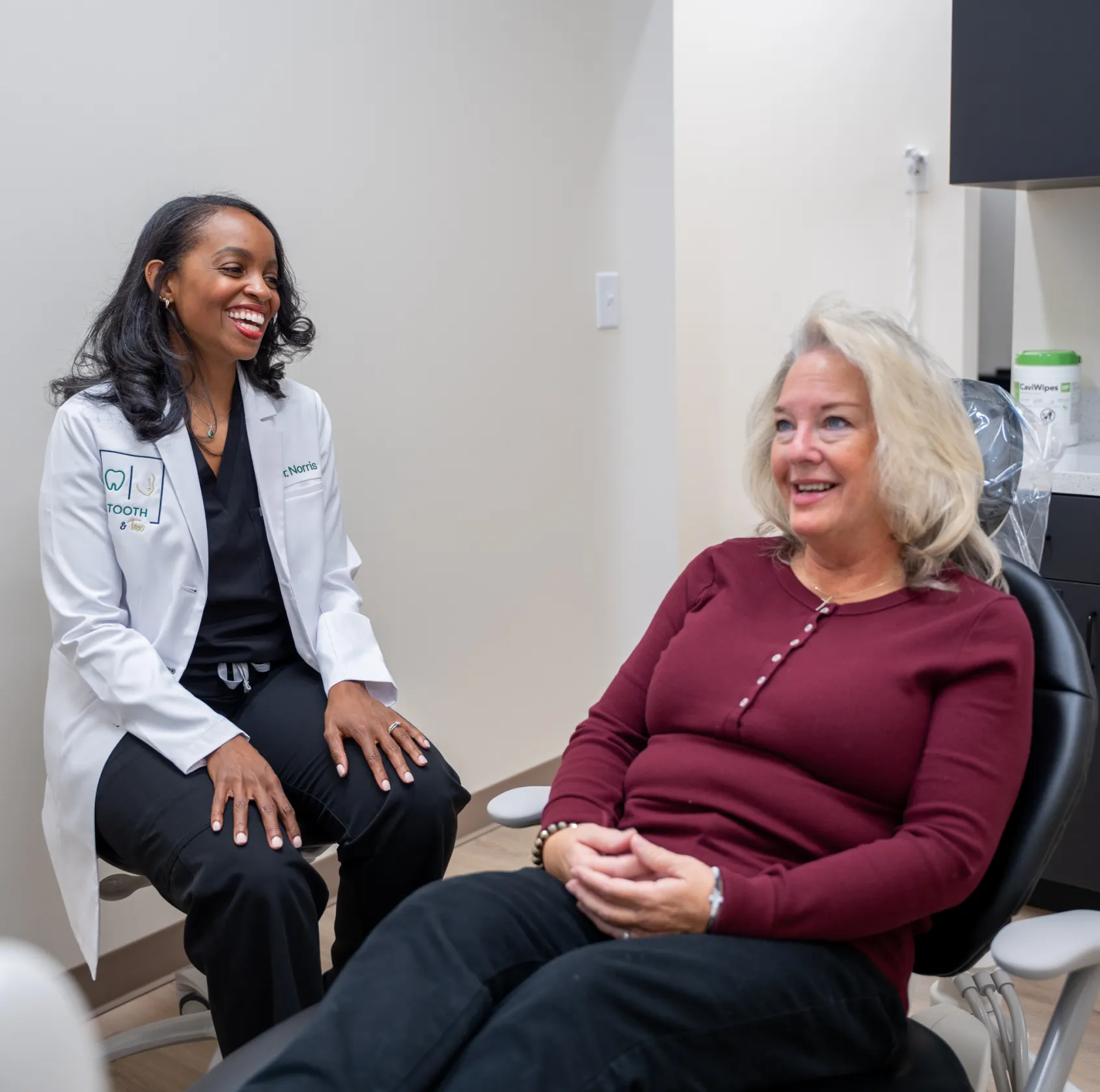 A female dentist in a white coat talks to a female patient sitting in a dental chair.