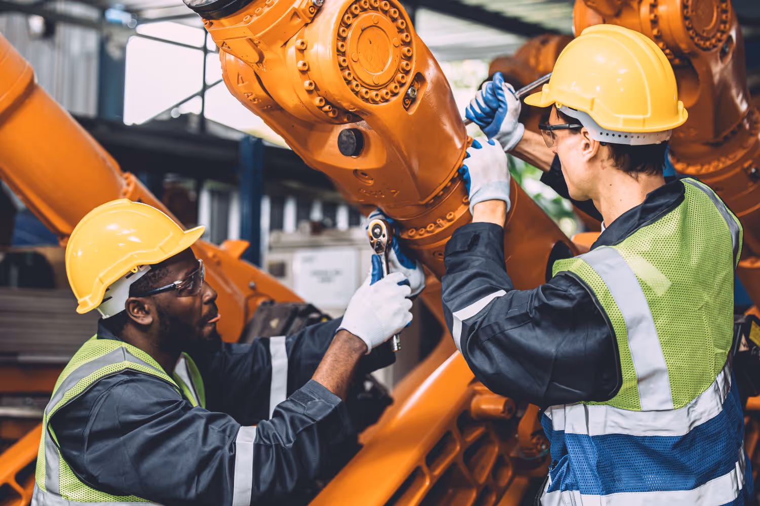 Twee technici met helmen en veiligheidsvesten onderhouden en repareren een grote oranje robotarm.