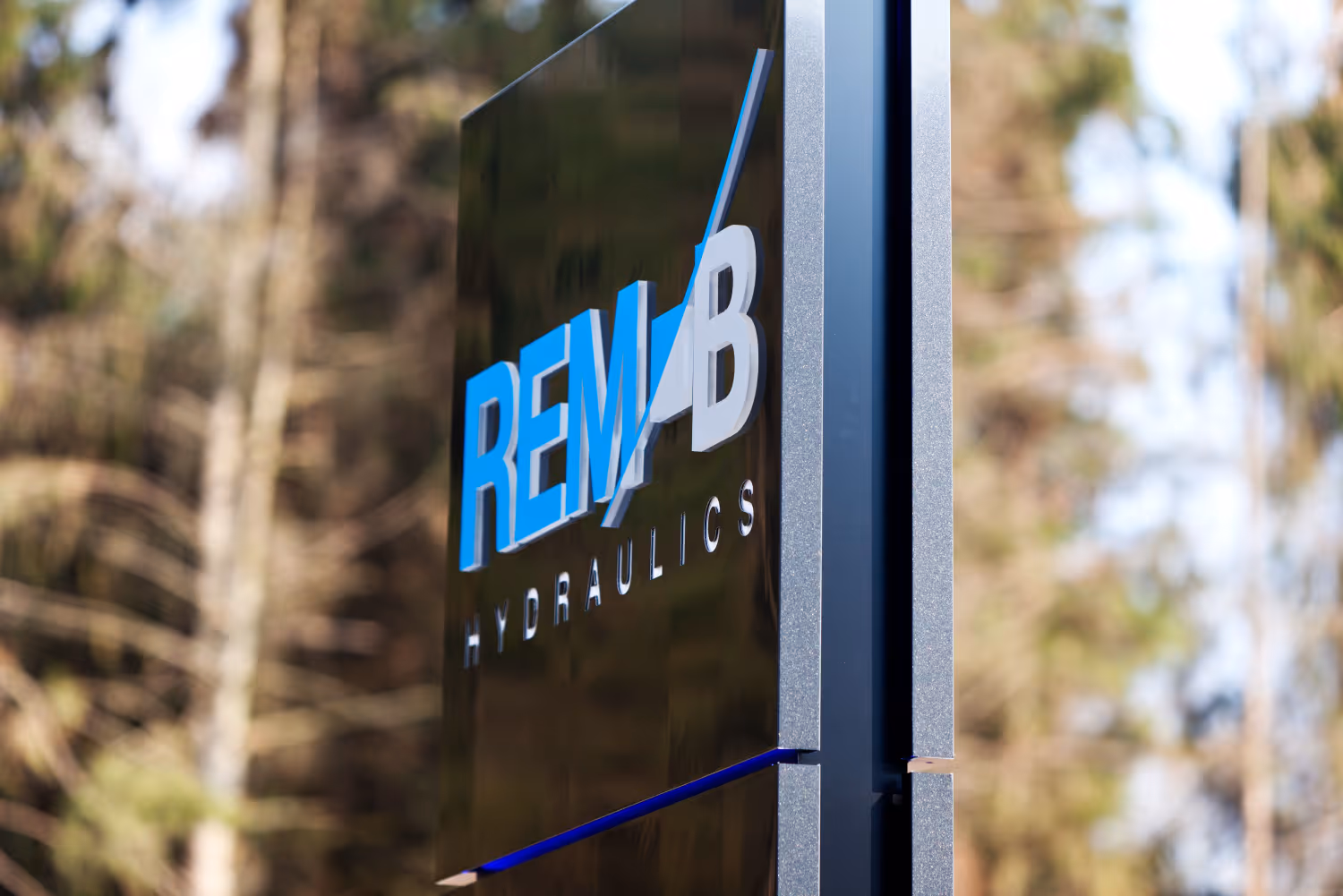 Bord met logo van REM B Hydraulics buiten, wazige bomen op de achtergrond.