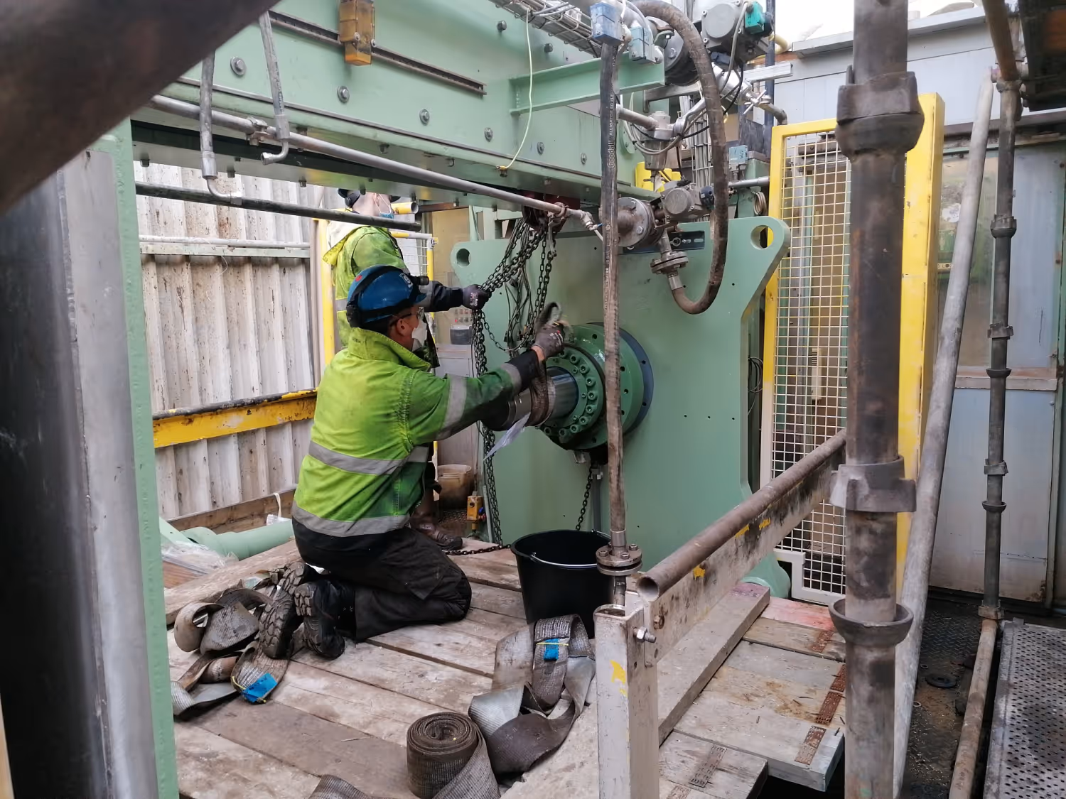 Twee arbeiders in veiligheidskleding werken aan een grote machine met kettingen op een bouwlocatie.
