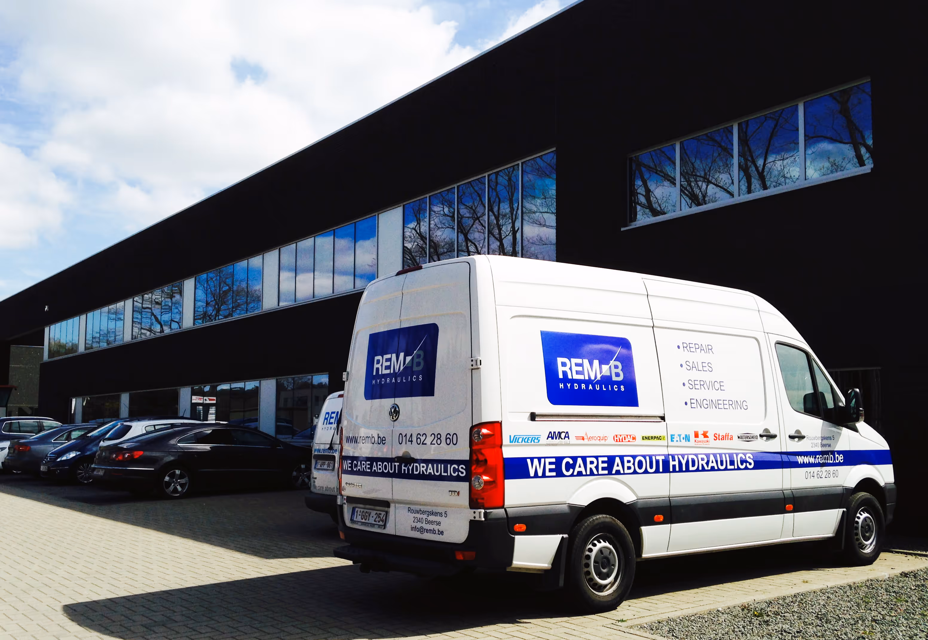 Wit REMB Hydraulics bus geparkeerd bij modern gebouw met reflecterende ramen.