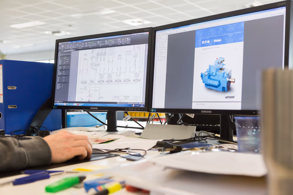 Persoon werkt met technische tekeningen en productafbeelding op dual monitors op een rommelige bureau.