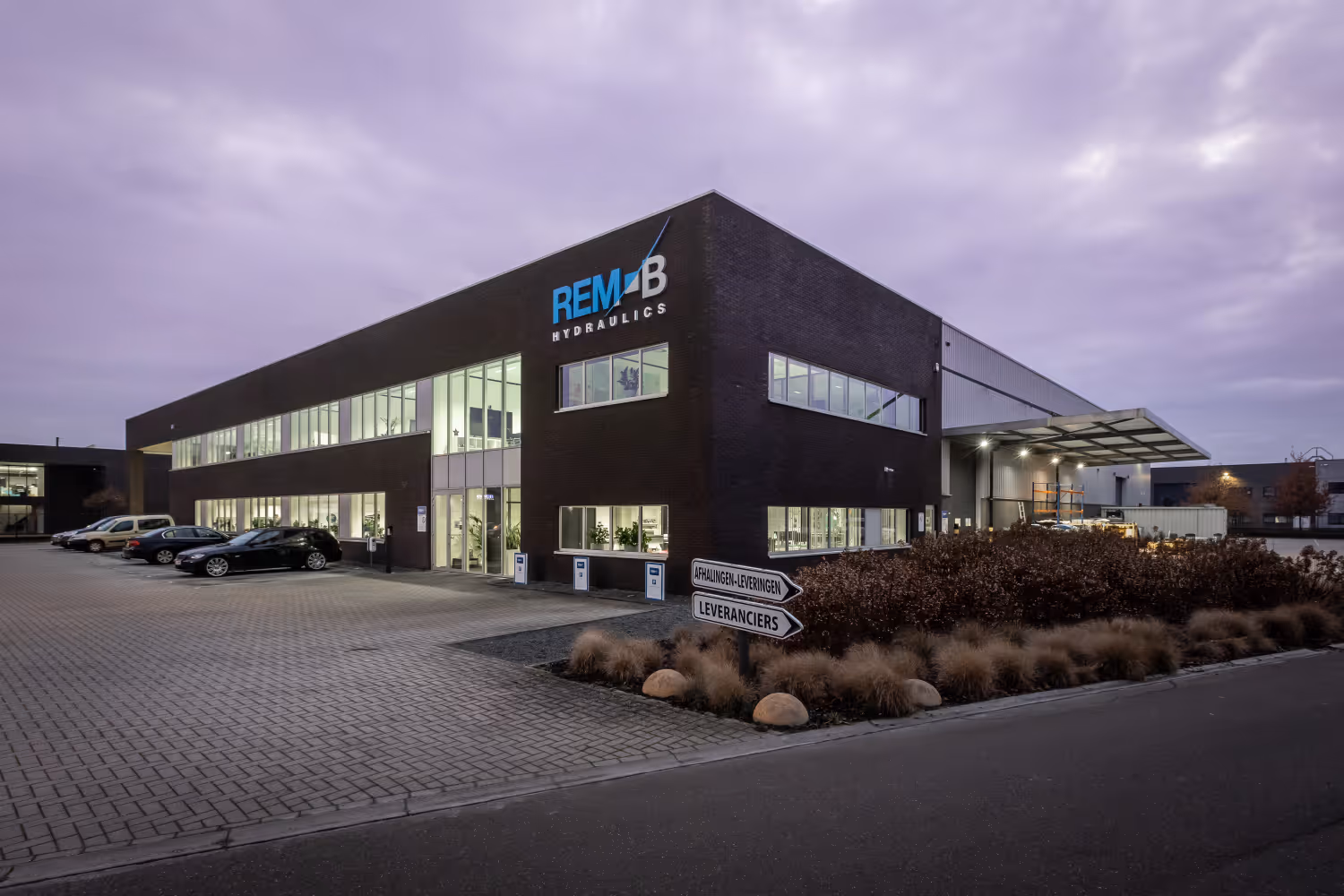 Modern REM-B Hydraulics building at dusk with parked cars and directional signs in front.