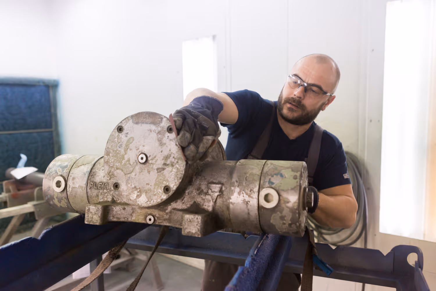 Man met veiligheidsbril en handschoenen werkt aan een grote versleten metalen machine in een werkplaats.