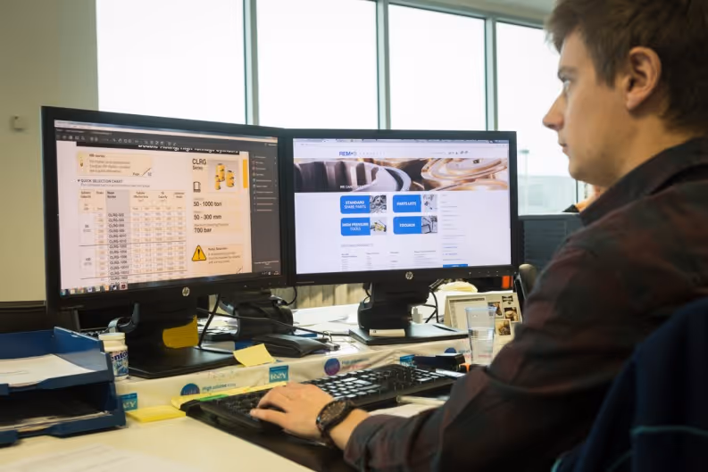 Man werkt op kantoor aan bureau met twee computerschermen en toetsenbord bij raam.