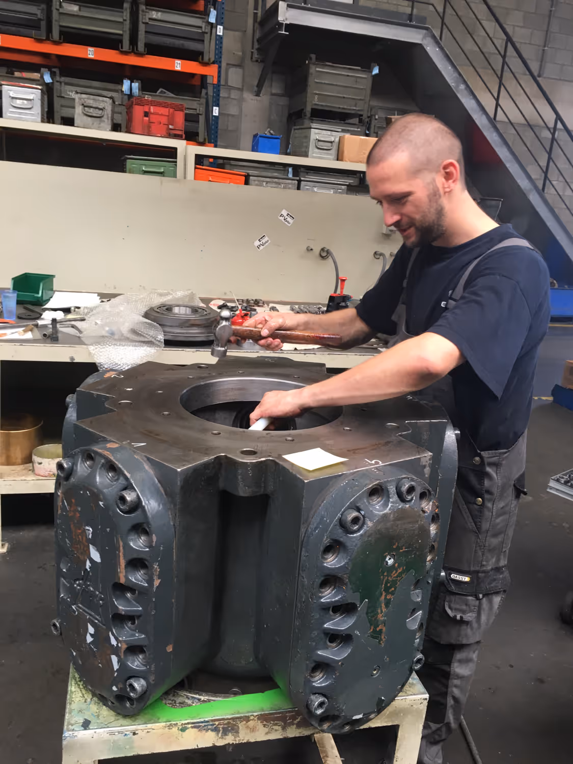 Man werkt aan grote metalen machineonderdeel met hamer in een werkplaats.